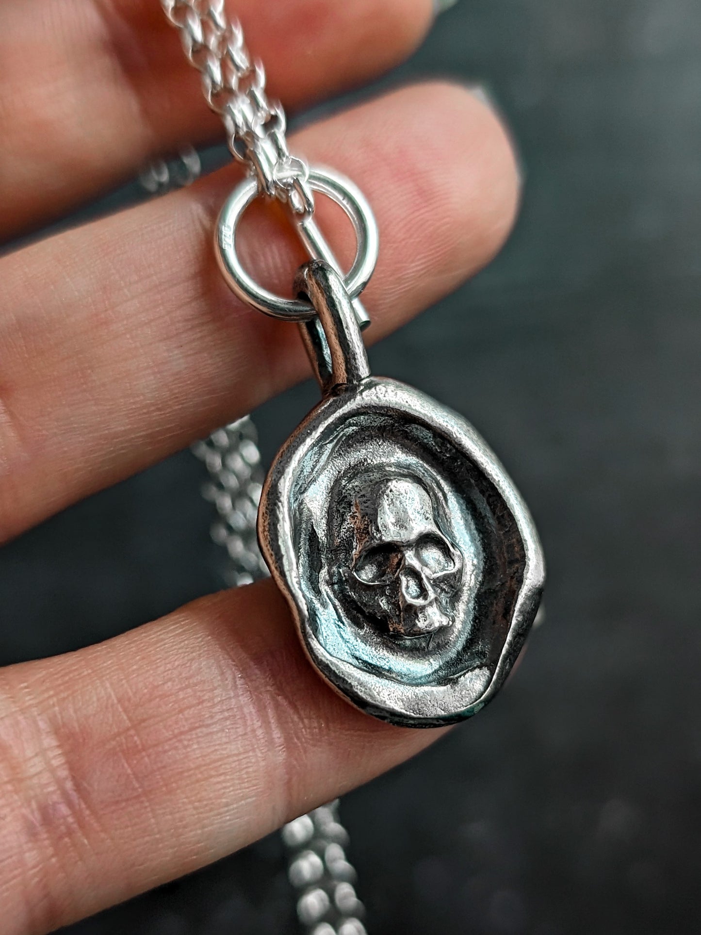 A darkened silver gothic skull pendant being held up-close by a hand to show the scale