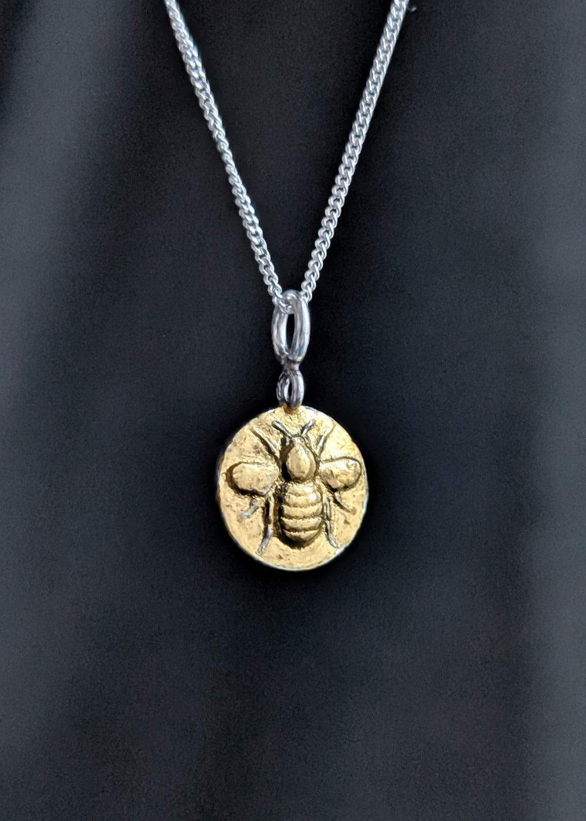 A round gold bee necklace hanging from a Sterling Silver chain in front of a dark, light-streaked background