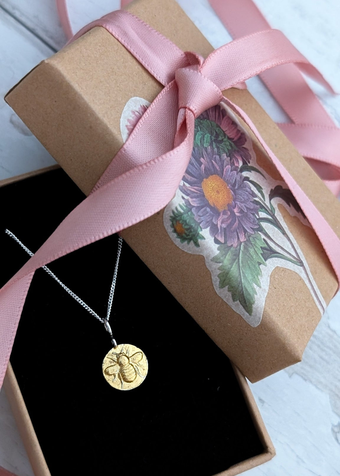 A tiny gold bumblebee circle pendant resting on black velvet padding in a floral gift box, tied with a pink box