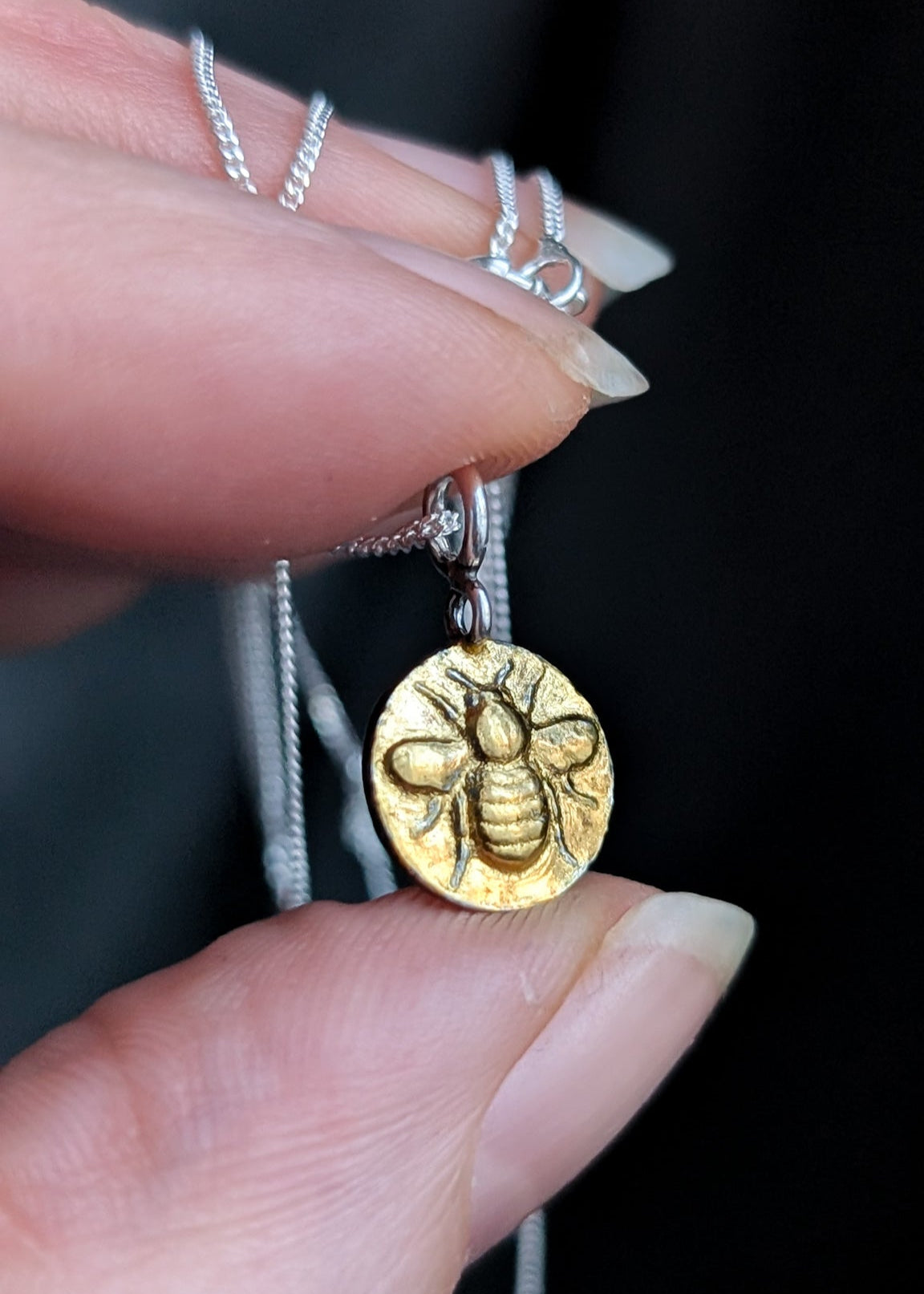 A shiny gold bee coin charm is held in a manicured hand, photographed close-up