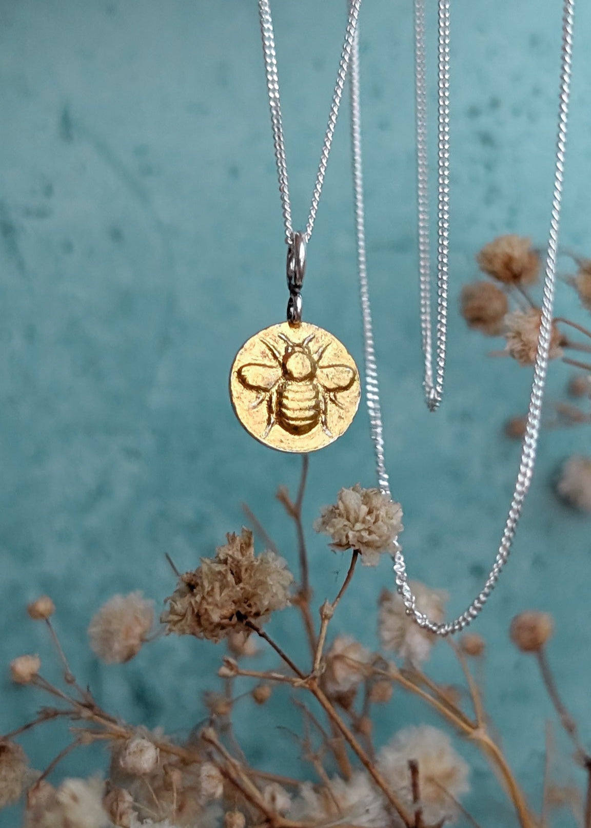 Petite gold bee medallion pendant hanging in front of a sky blue background with whispy summer florals bordering the image