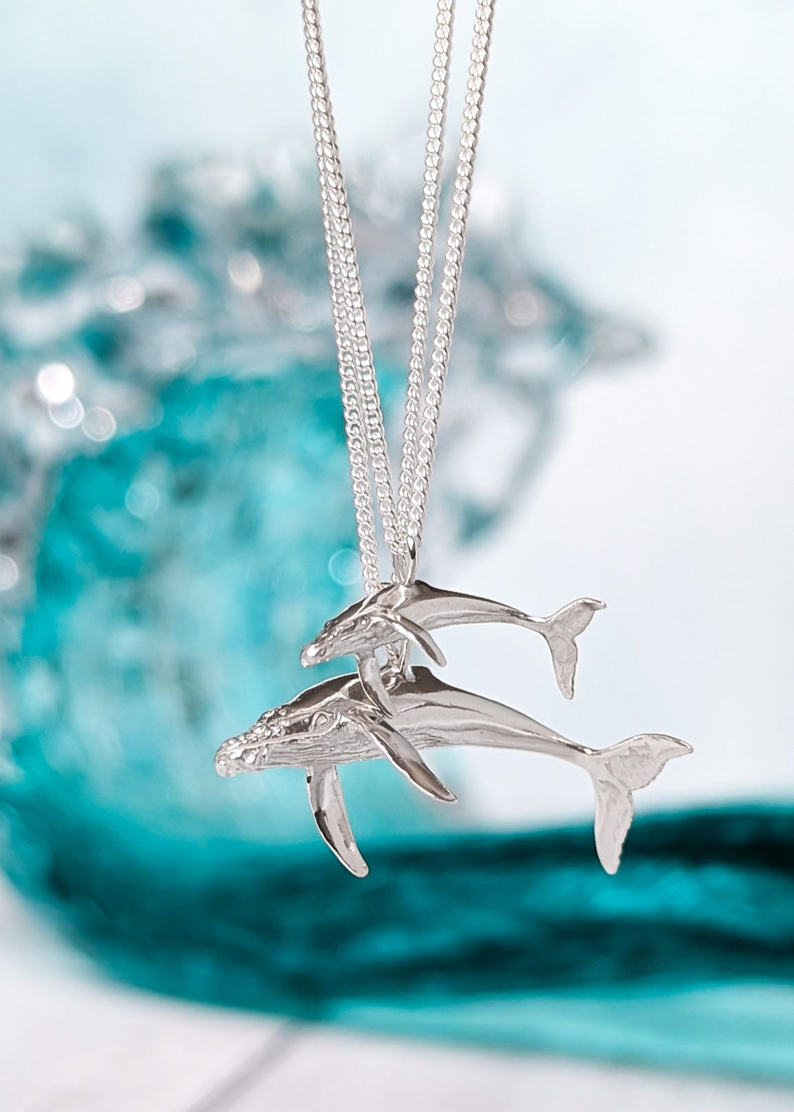 Identical silver whale charms of different sizes, the smaller pictured above, pictured in front of a crashing blue ocean wave.