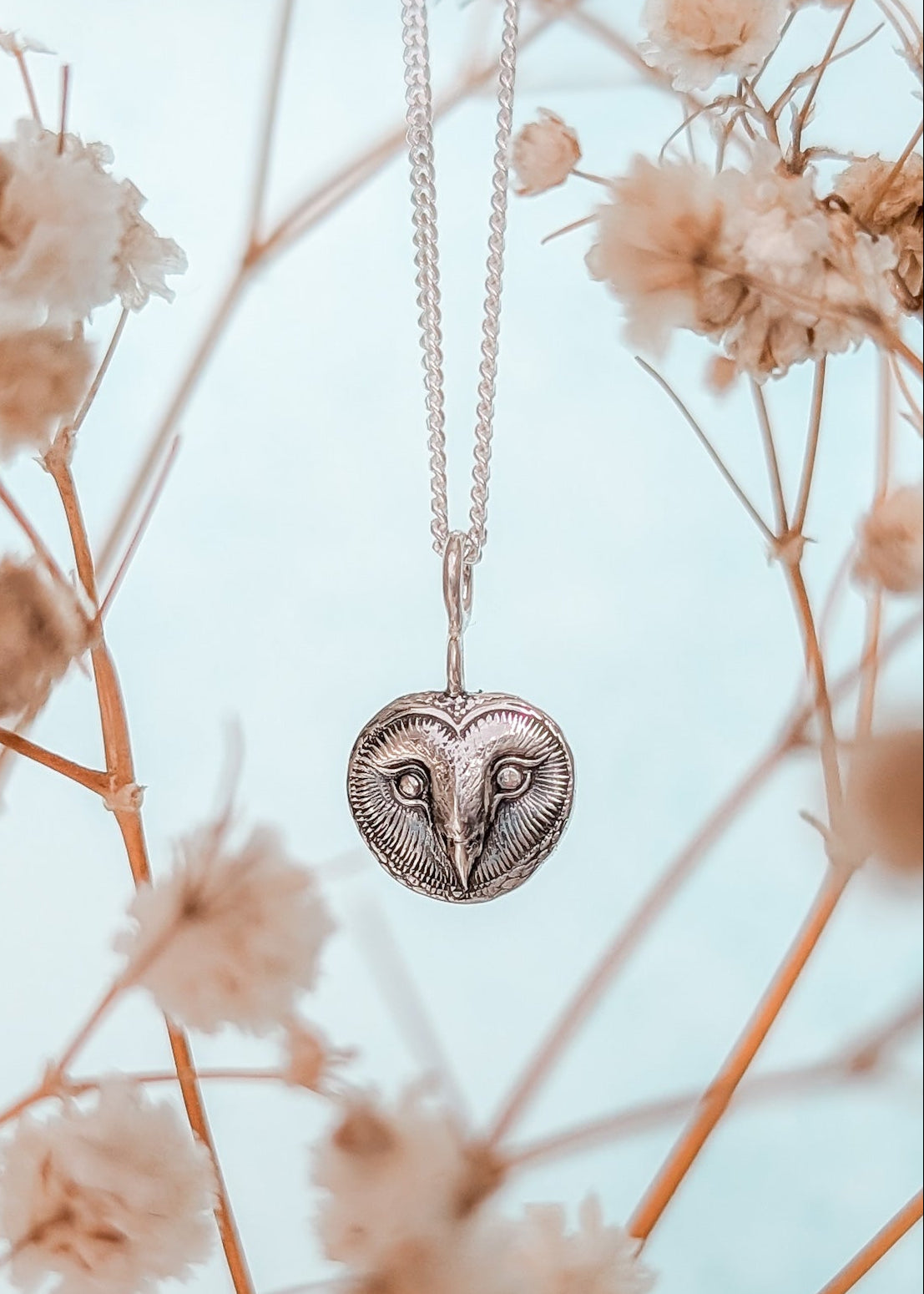 Heart-shaped owl face pendant necklace hanging among dried flowers with a light blue background