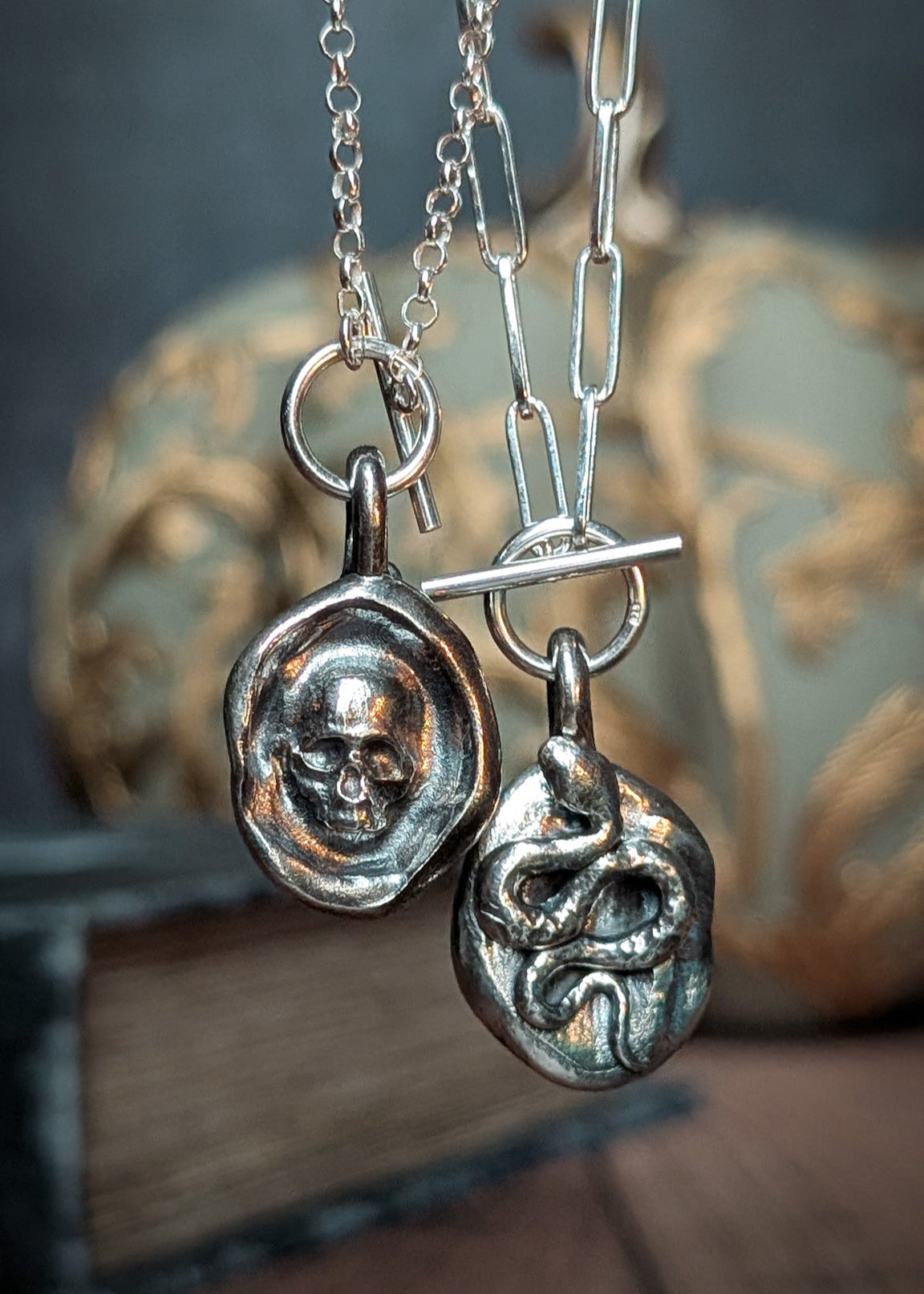 A silver wax seal style skull pendant hanging beside the reverse of the same pendant which features a coiled serpent