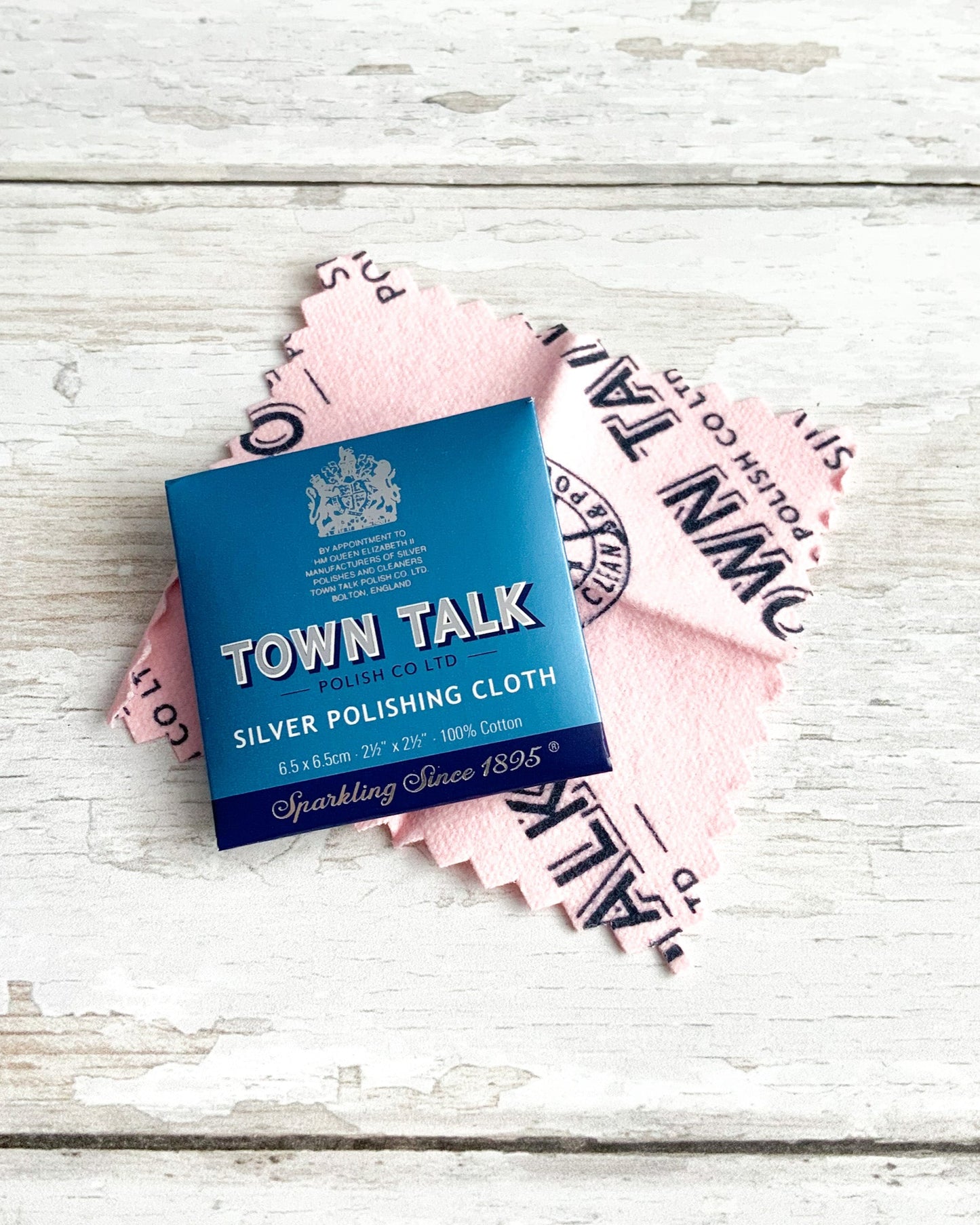 A pale pink suede textured polishing cloth for silver jewellery, pictured on a white wooden surface beside its blue envelope packaging