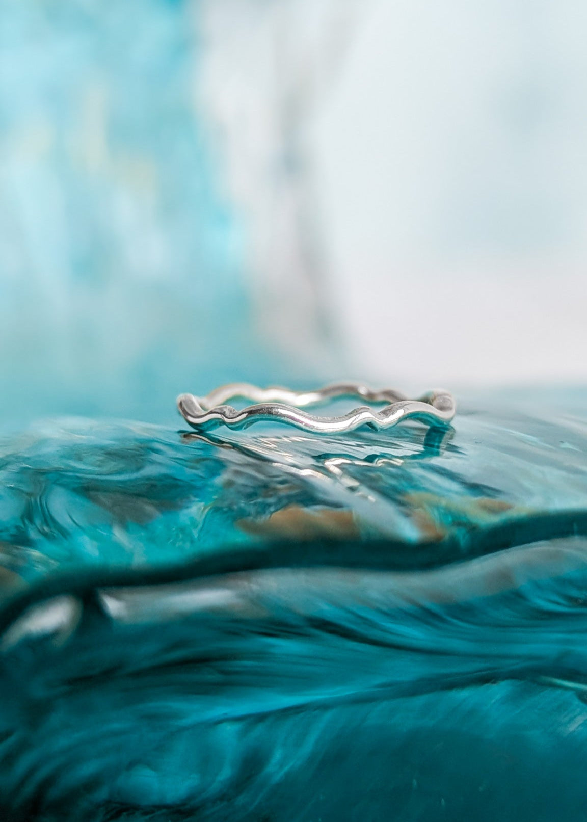 A skinny silver wave ring pictured on top of ocean blue glass that replicates the sea