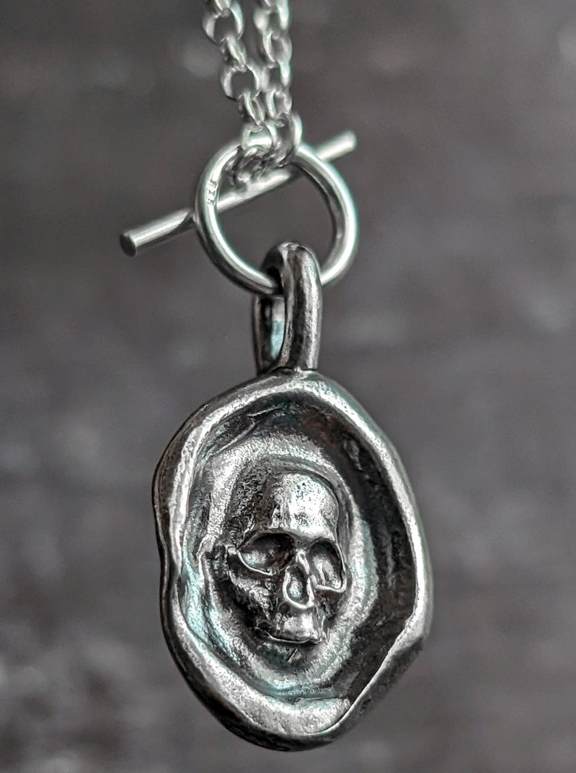A memento mori wax seal style skull jewellery charm hanging from a T-bar chain