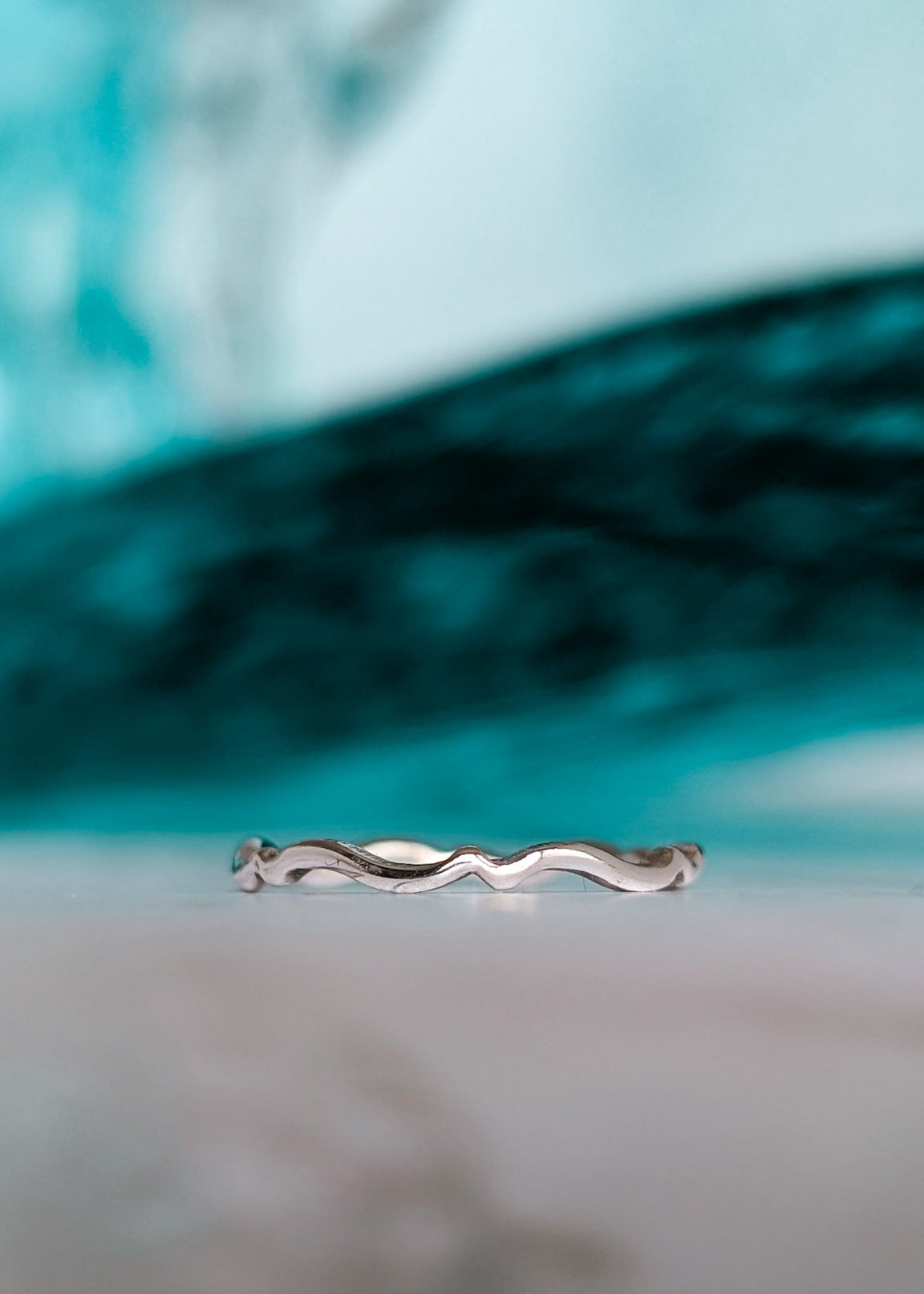 A shiny silver scalloped wave ring pictured in front of a tuquoise wave