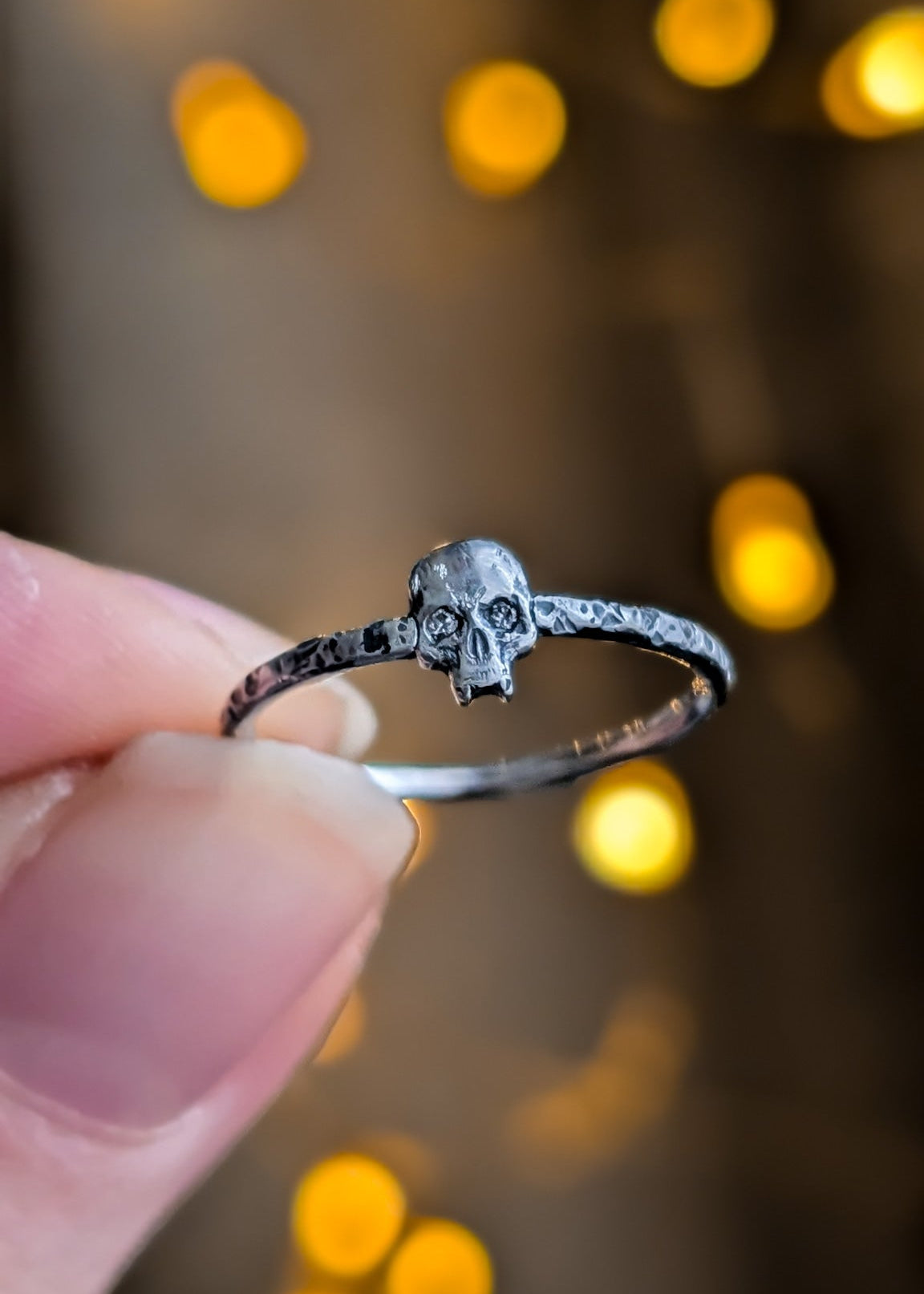 Close-up of fingers holding a silver skull ring with diamond eyes against a warm, blurred background with yellow bokeh lights.