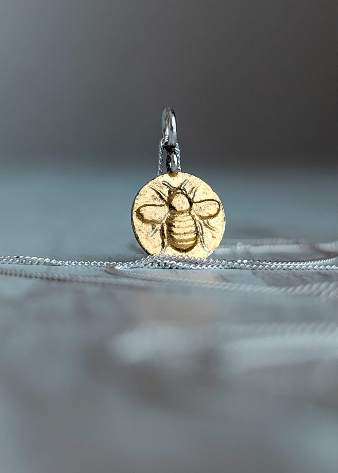 A yellow gold bee necklace,  round, resting on silver chain hanging from its bail. Photographed on a soft grey background