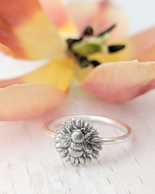 Silver ring with a sunflower and bumblebee design, resting on a white wooden surface with a blurred flower in the background