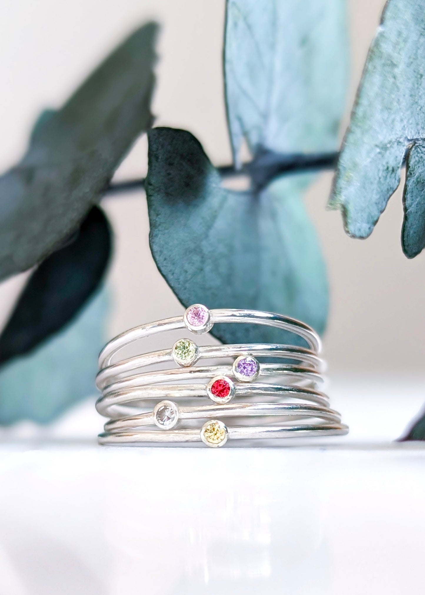 Pink, green, purple, red, clear and yellow stone solitaire rings all stacked together in front of some eucalyptus foliage