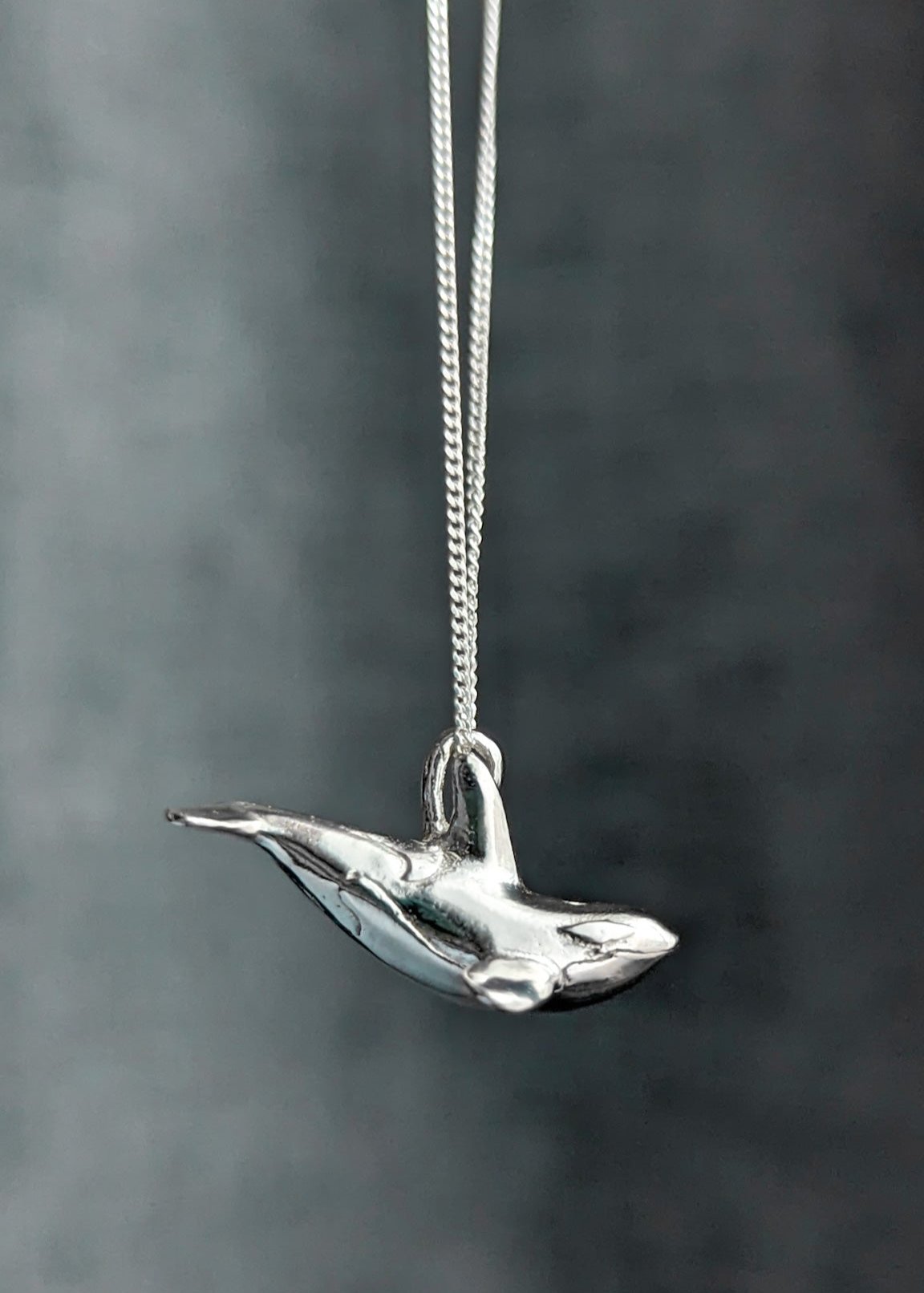 A realistic silver orca charm hanging from a chain against a textured cool grey background