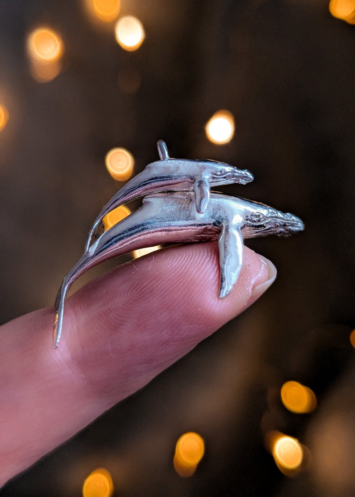 Mum and baby humpback whale charms resting on a fingertip in front of a warm bokeh lighting backdrop