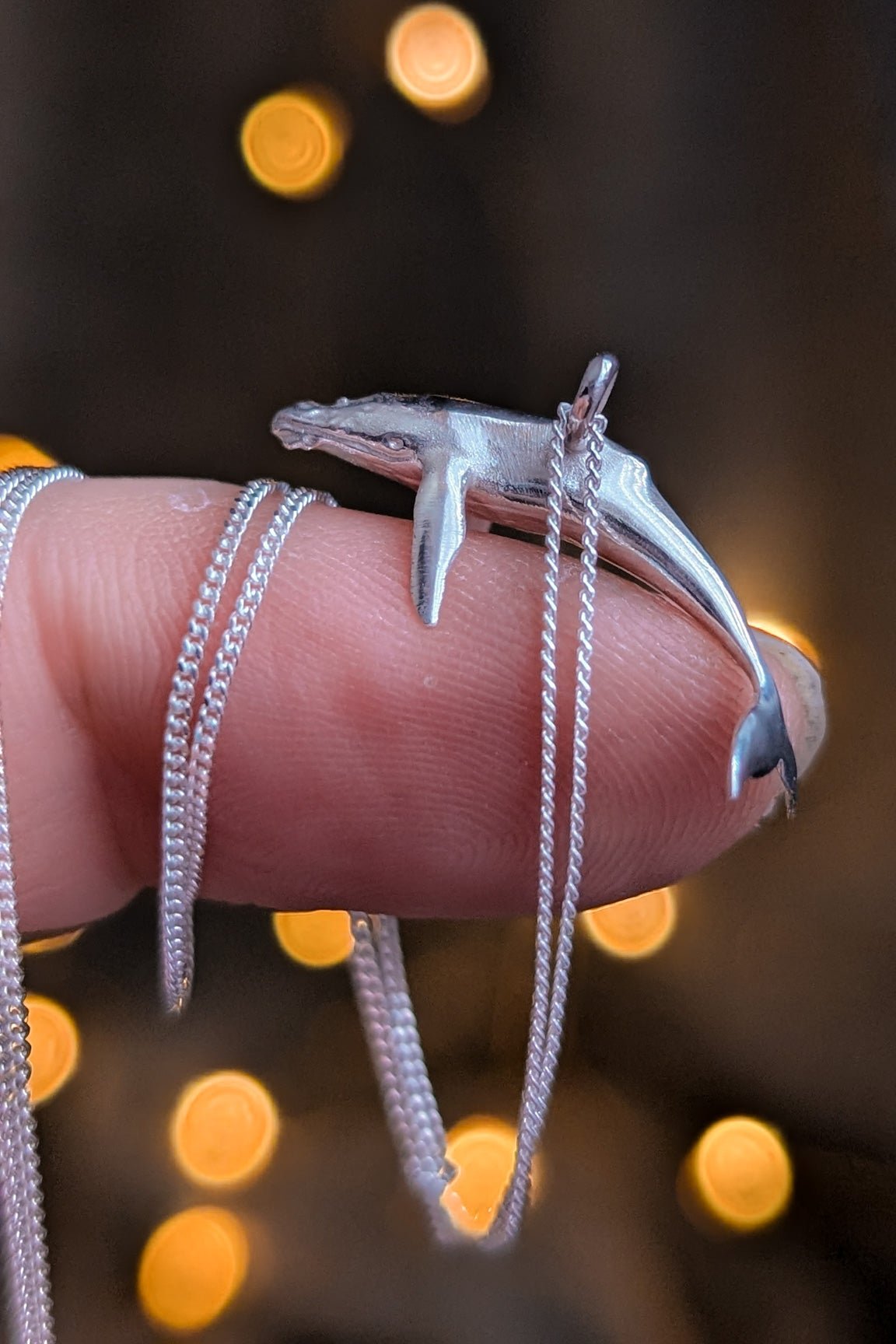 Shiny silver miniature whale pendant resting on a fingertip in front of a warm bokeh lit background