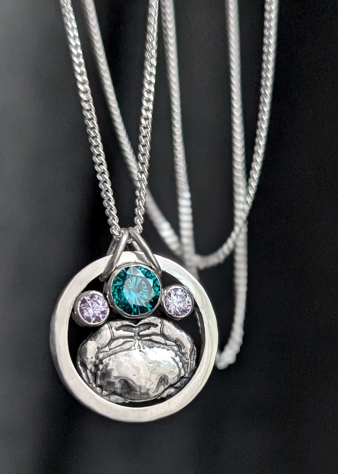 Silver circle necklace featuring a detailed silver crab, green blue gemstone and smaller pale purple stones, photographed on a dark background