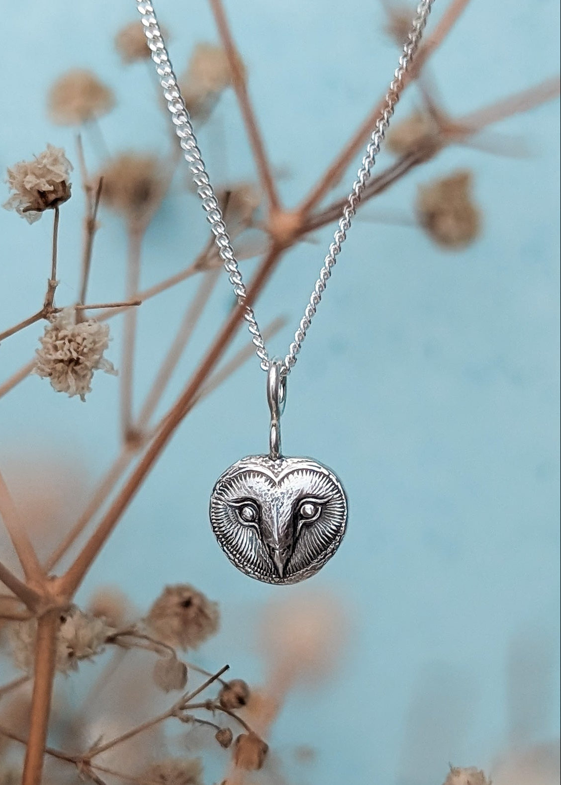 A detailed silver owl face pendant against a pale blue background with soft focus dried flowers