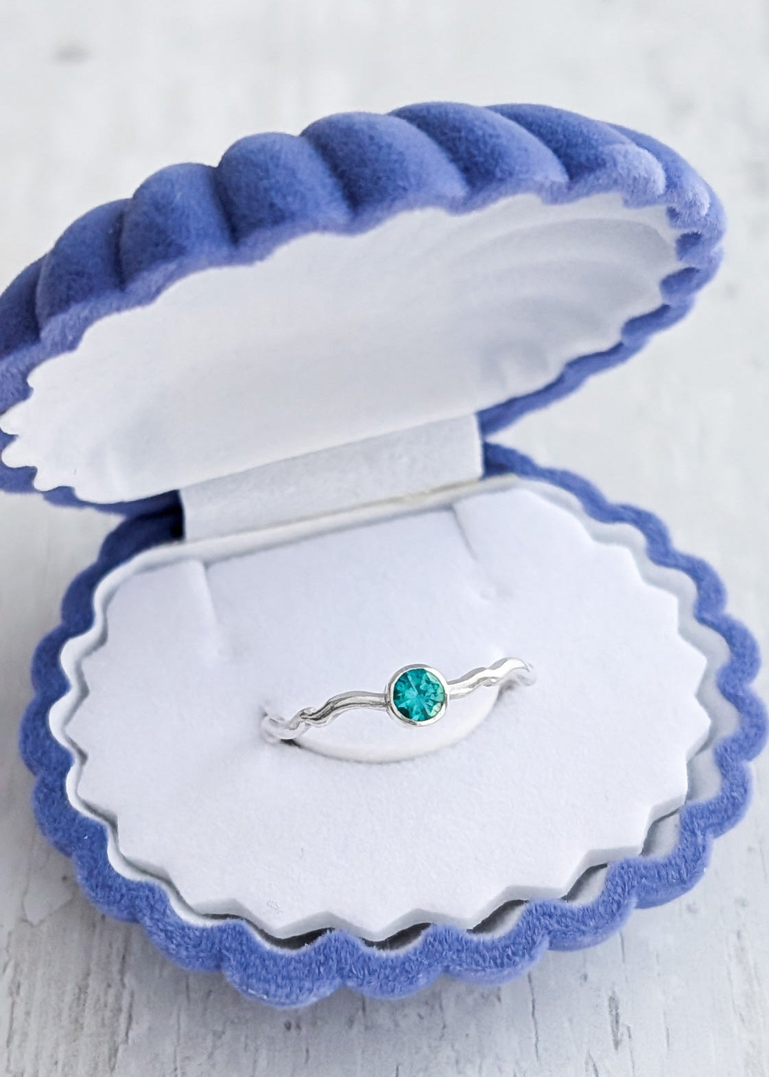 Silver wiggle wave ring with a blue-green gemstone displayed in an open blue shell velvet gift box on a light wooden surface.