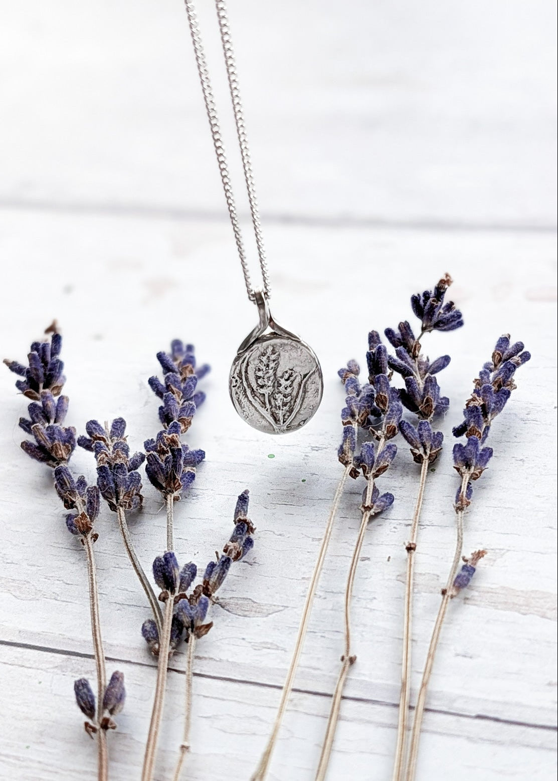 Textured silver oval pendant suspended from a silver chain above a wooden surface strewn with lavender flowers, the oval charm also features lavender flowers embossed in silver