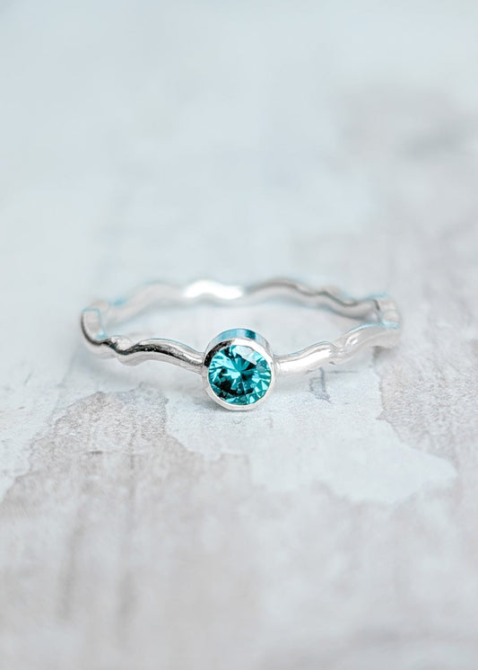A crimped wave pattern silver ring with an ocean blue gemstone resting on a white wooden surface