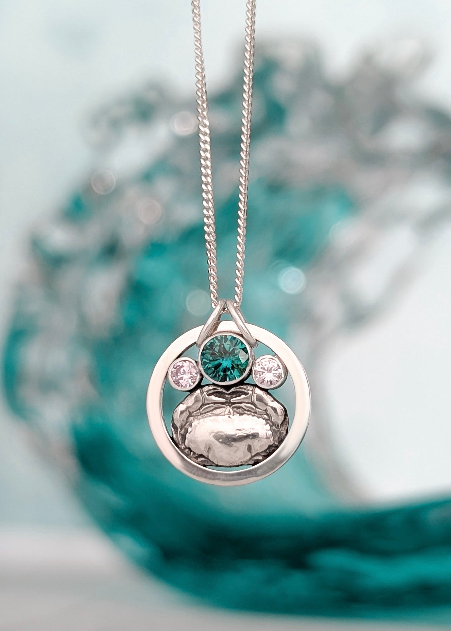 A shiny silver circle pendant with a shore crab silver charm inside and 3 round stones.  The centre stone is larger and ocean blue-green.  The necklace is photographed in front of a cresting turquoise wave.