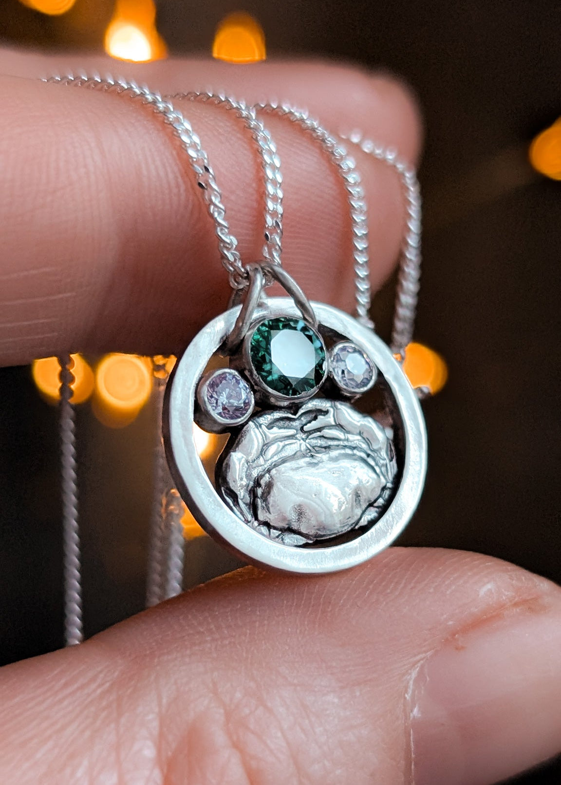 Silver circle necklace featuring a crab design held between fingers against a blurred bokeh lights background.