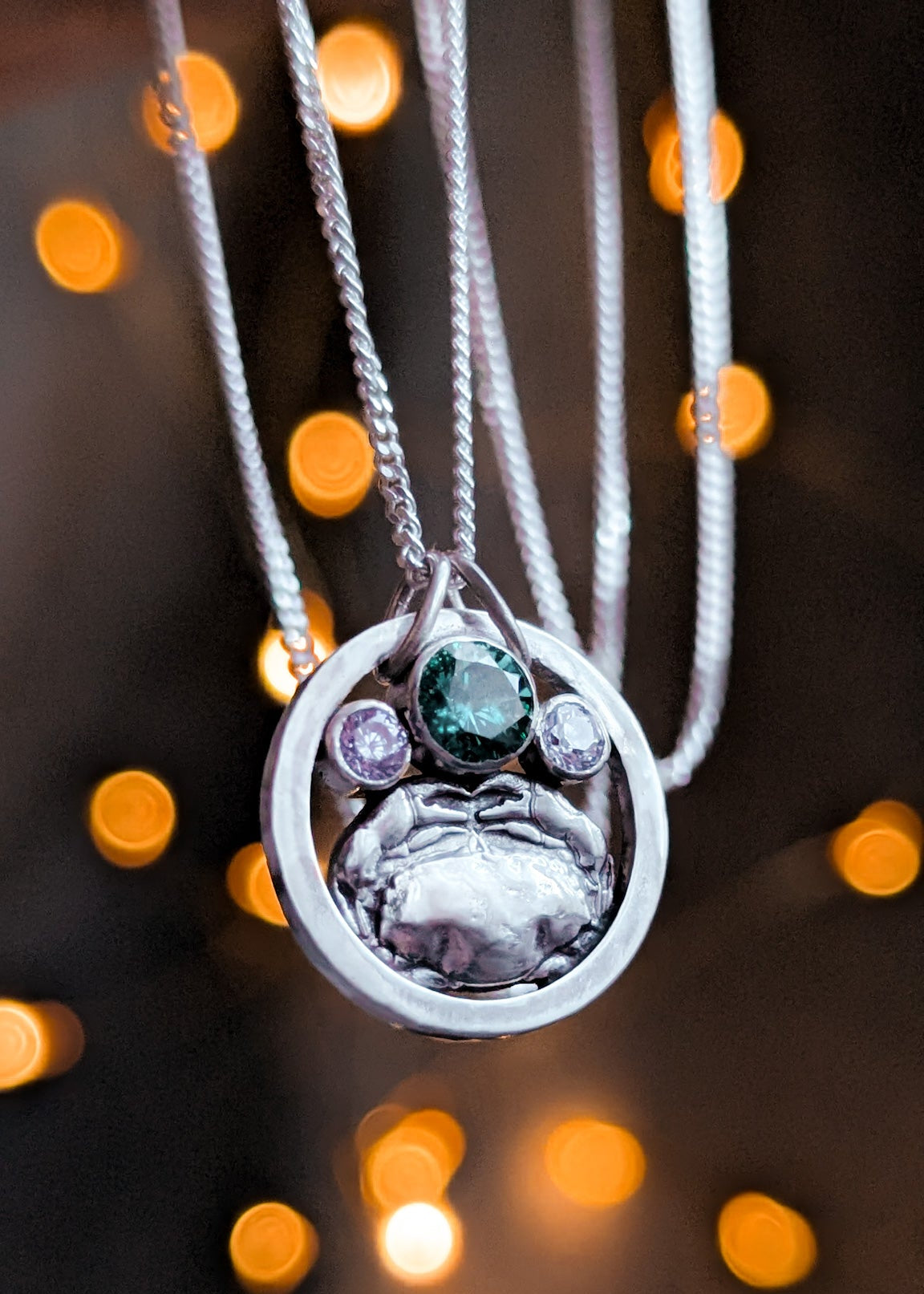 Silver circle pendant with a detailed silver crab and colourful gemstones against a blurred bokeh light background