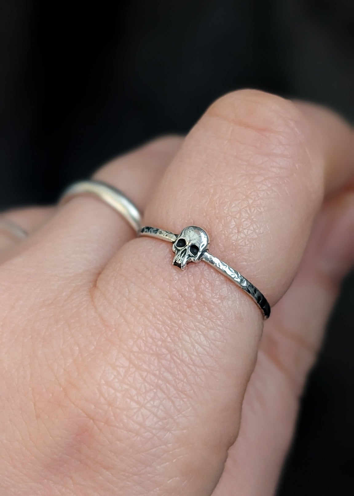 Close-up of a hand in front of a dark background wearing silver rings, in focus is a small skull designs on the index finger