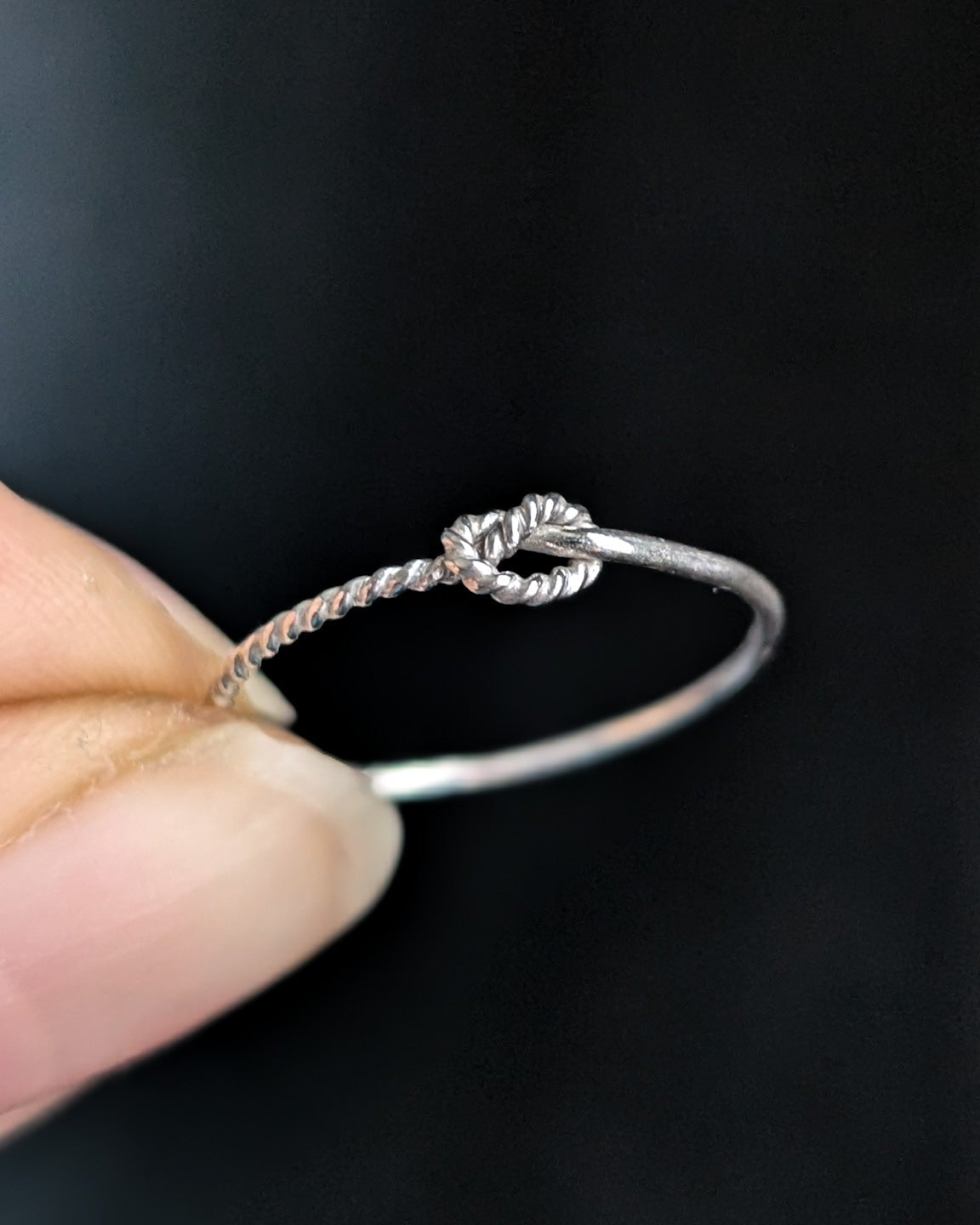 Close-up of a hand holding a delicate silver ring with a knotted rope detail in front of a dark background.