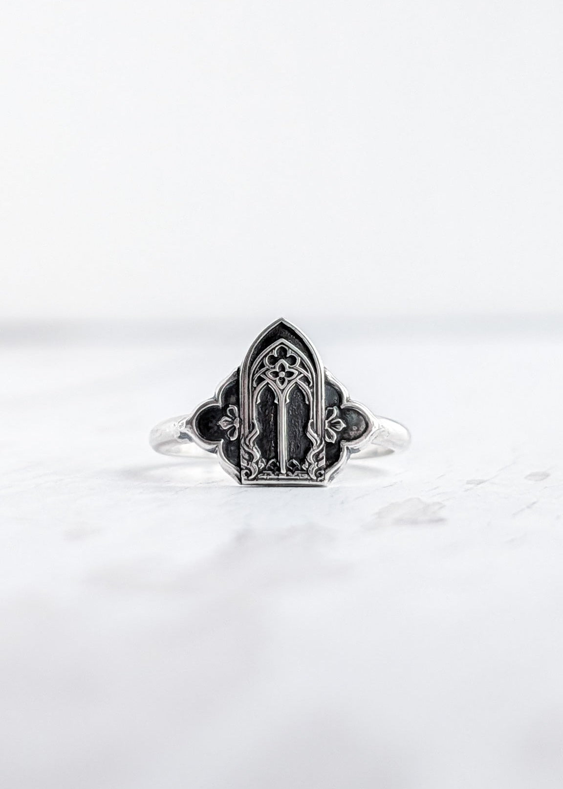 A two tone silver and darkened silver cathedral window ring rests on a lightly textured white wooden surface
