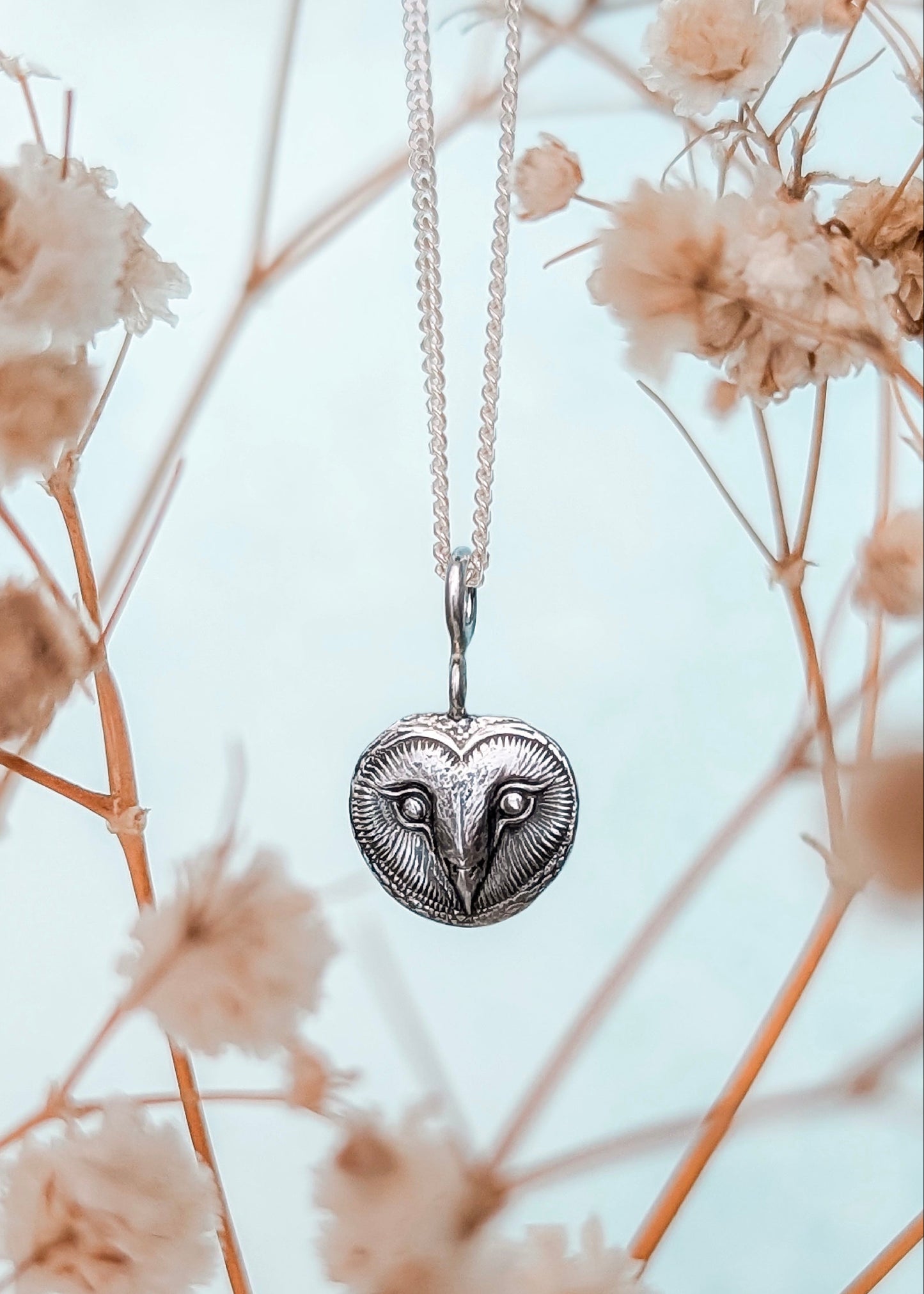 Silver heart-shaped owl face pendant necklace hanging among dried flowers with a soft focus background