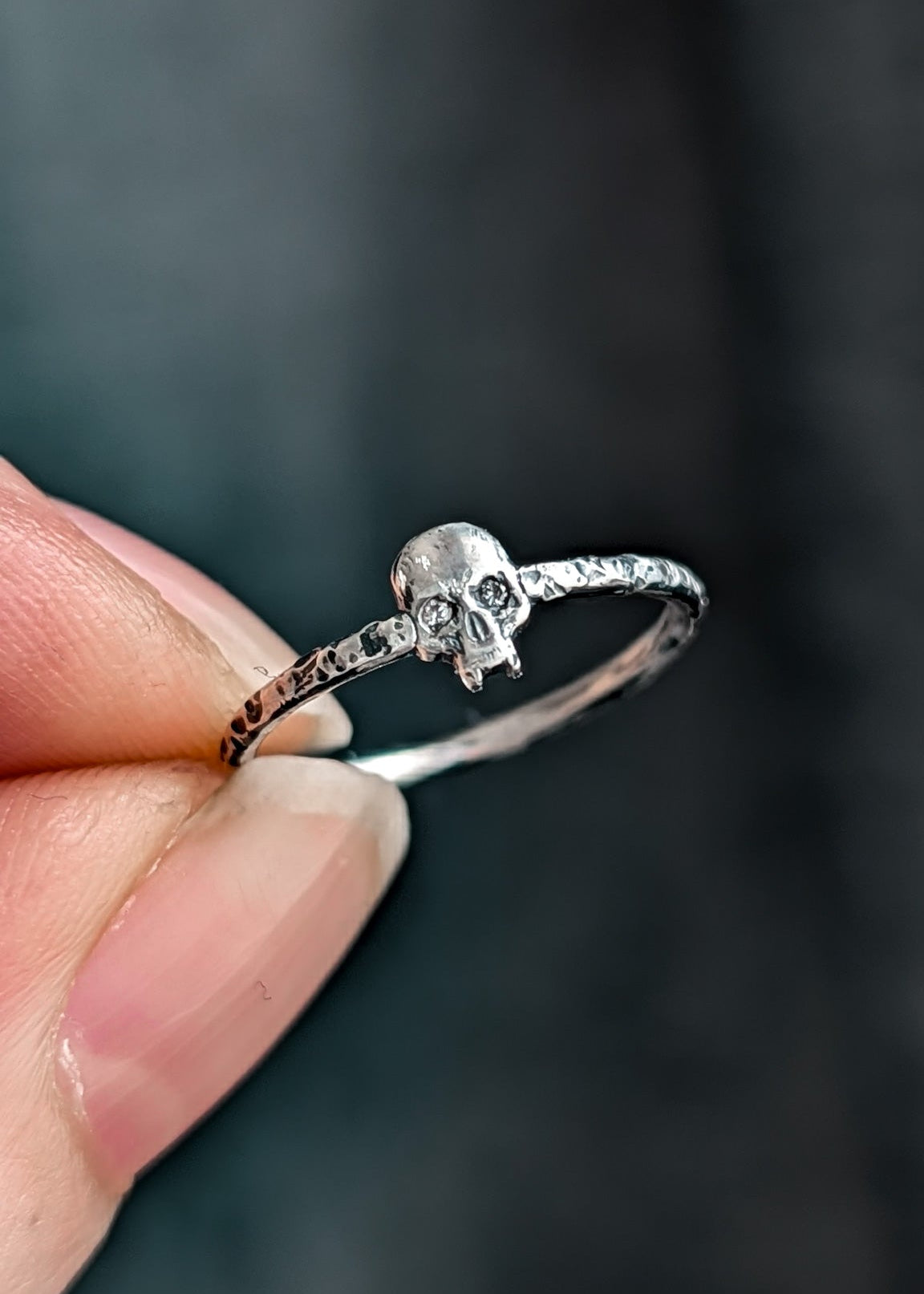 Close-up of a silver skull ring with diamond set eyes held between fingers against a dark, blurred background.