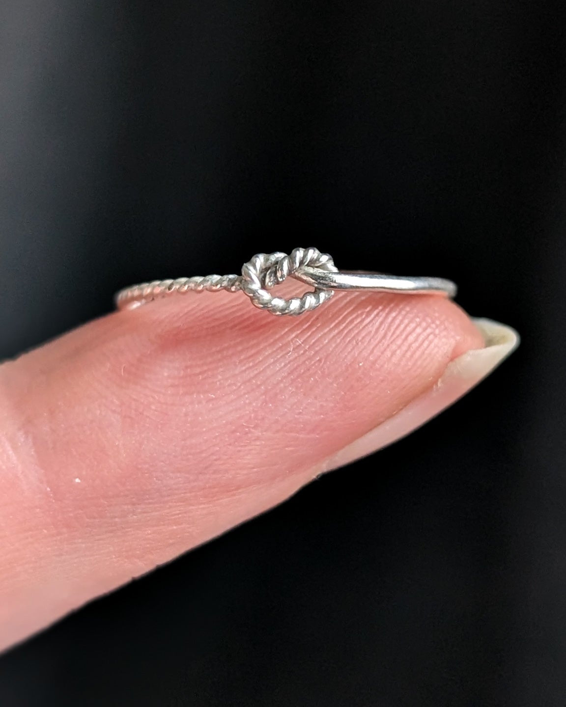A delicate silver ring with twisted rope wire and a knot design is held on a person's fingertip against a dark background.