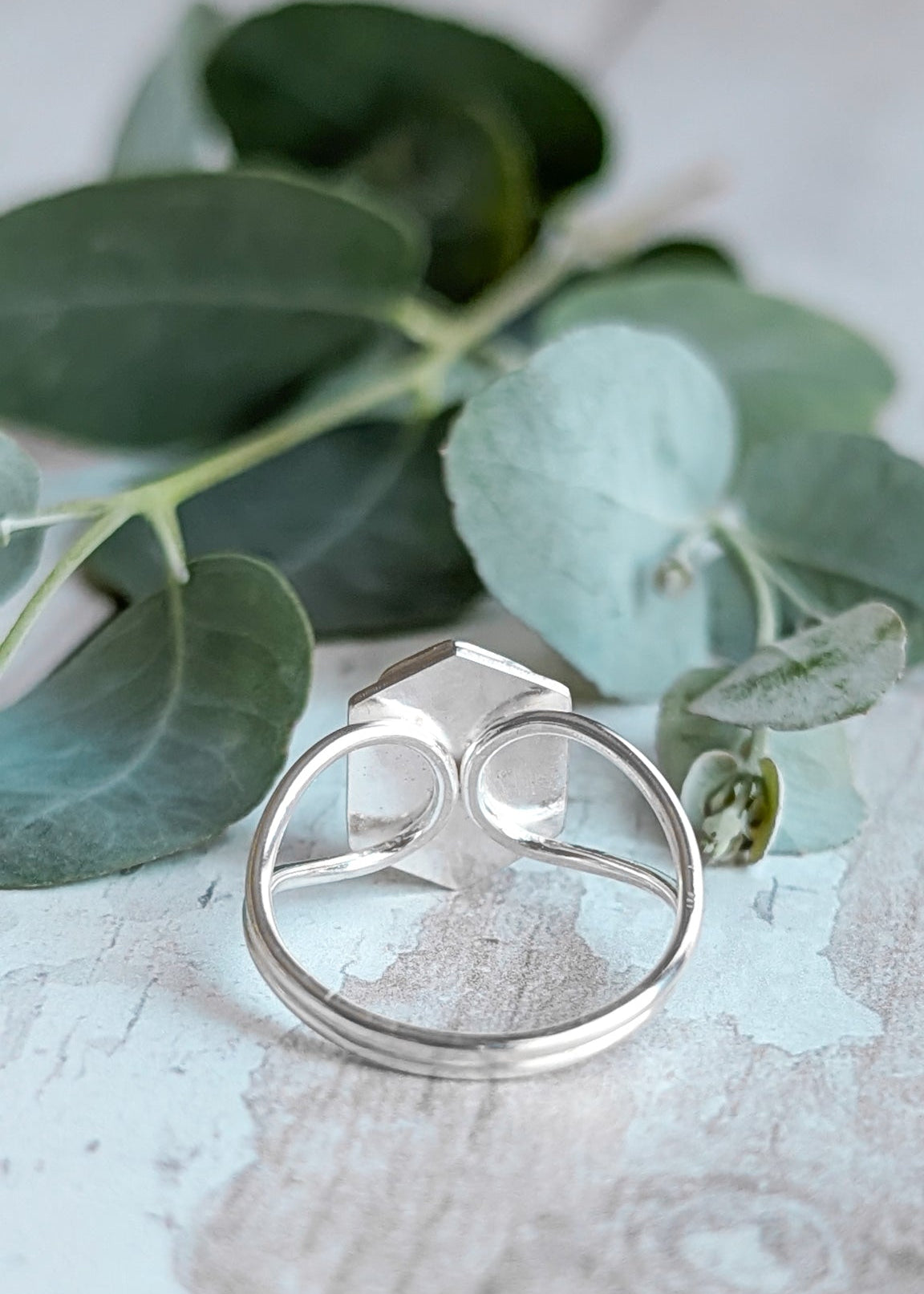 A silver ring rests on a light wooden surface facing away from the viewer to show the underside, decorative foliage rests just beyond it