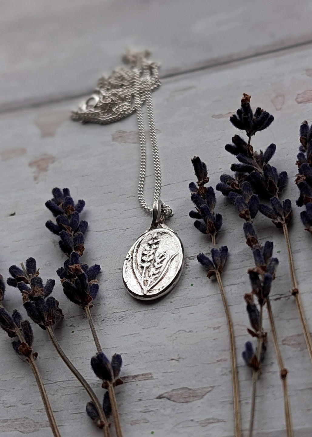 A slightly darkened scene showing a shining silver oval pendant amongst lavender flowers, with a silver chain flowing behind the pendant charm across a white wooden surface