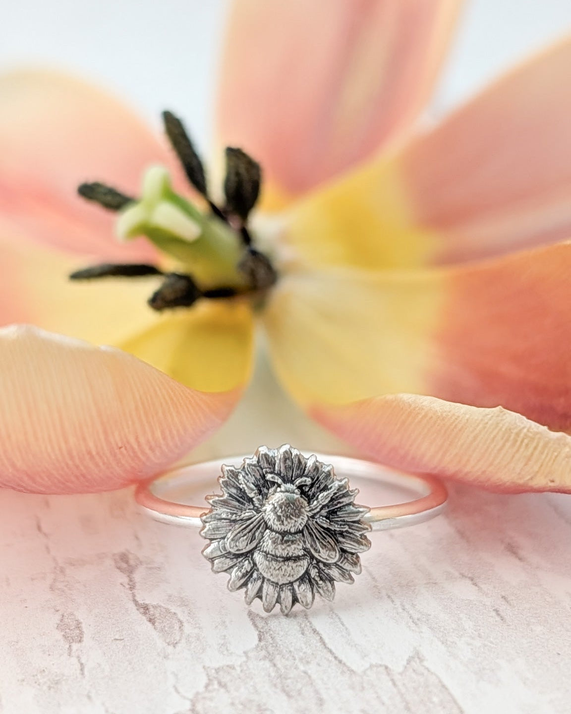Silver ring with floral bumblebee design on a white wooden surface with a flower in the background