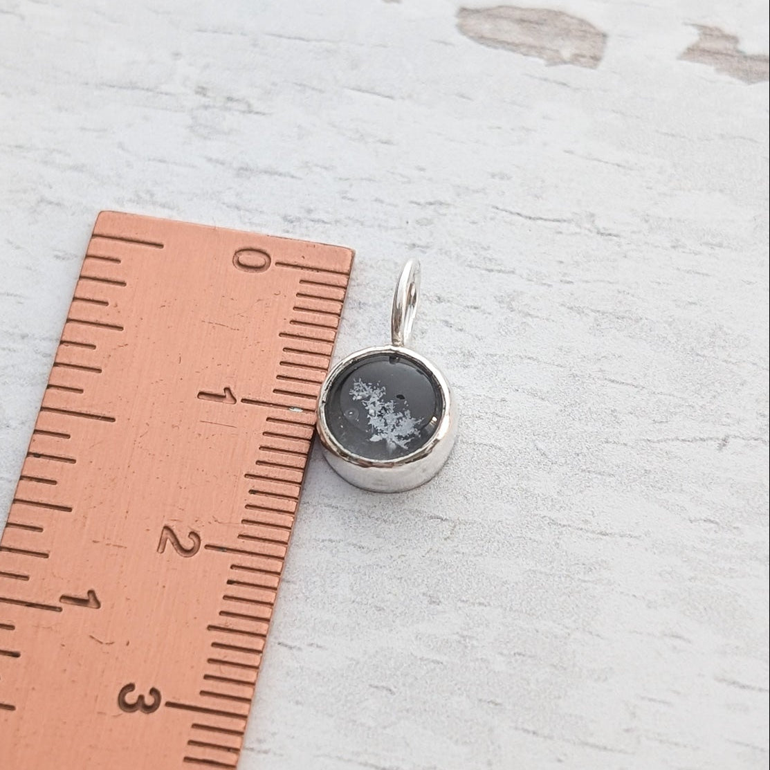 Small black and silver pendant containing a real preserved snowflake, pictured on a white wooden surface beside a ruler for scale