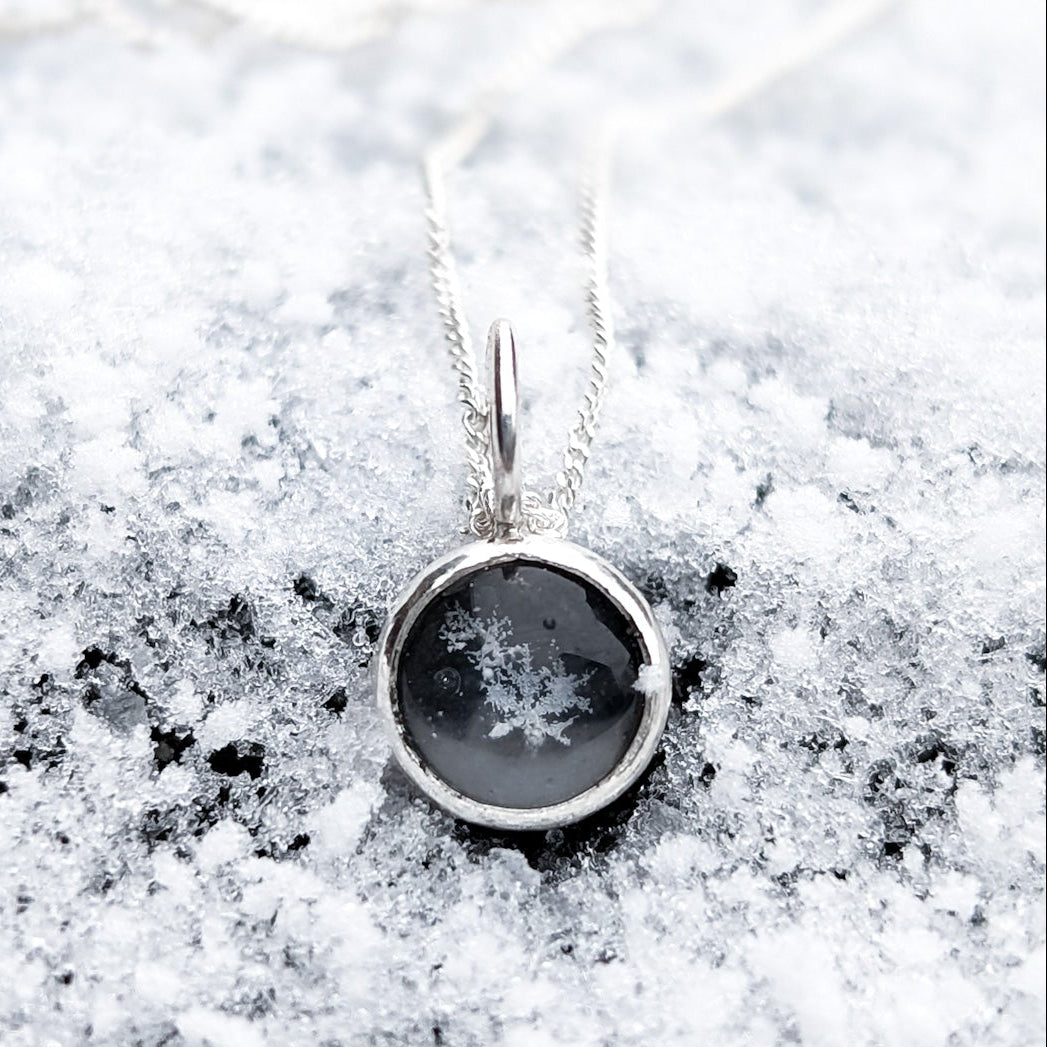 A round black stone pendant with a cluster of genuine preserved snowflakes in the middle, photographed on fresh snow