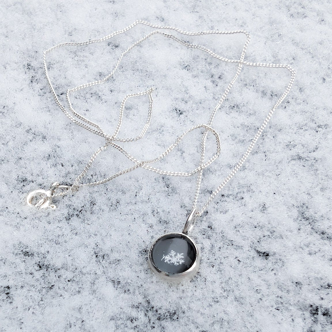 A small domed pendant on a silver chain resting in the snow.  The pendant charm is round, domed and holds a tiny white snowflake.