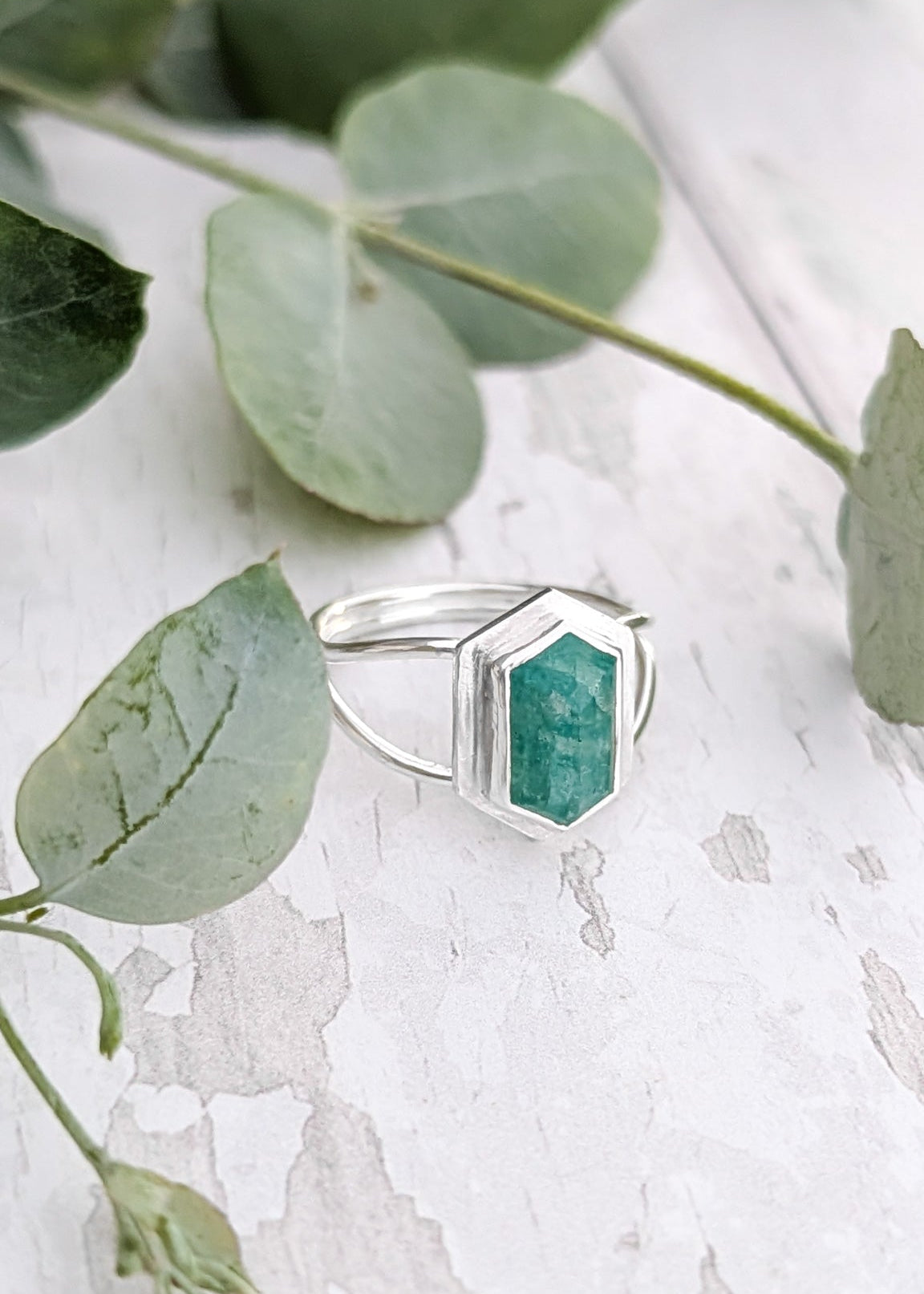 A silver Amazonite gemstone ring rests at a slight angle on a white wood surface beside some decorative eucalyptus