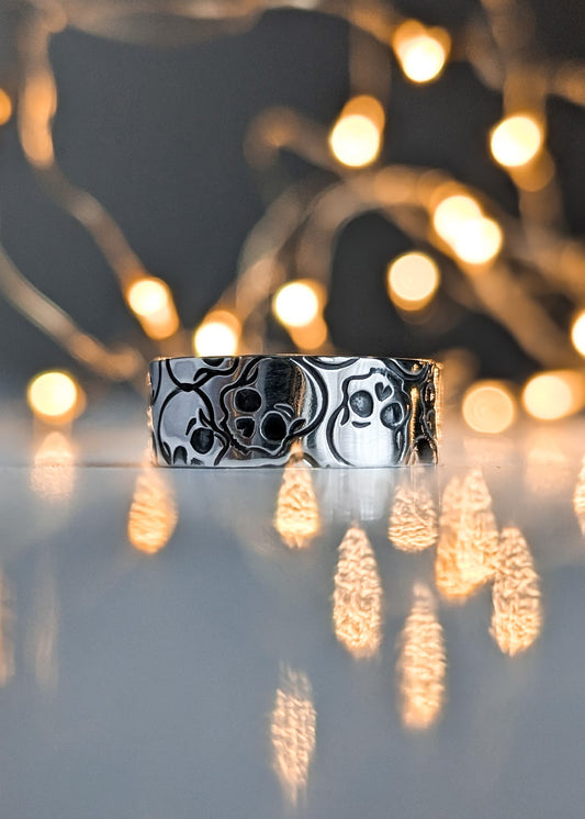 Silver ring with black skull pattern, pictured on a reflective surface with blurred lights in the background