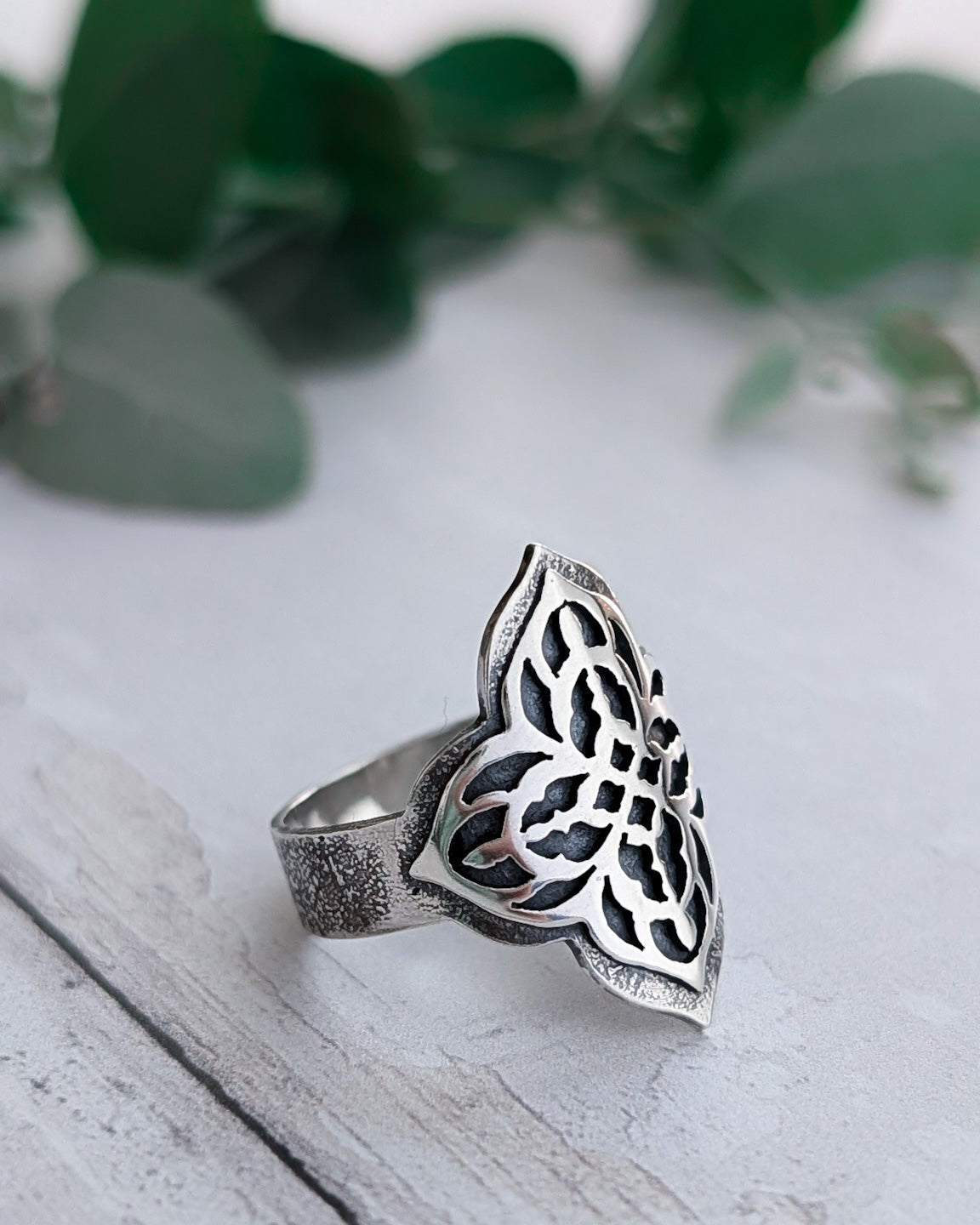 Side view of a textured and darkened silver quatrefoil signet ring with cathedral window cut-out detail front adornment.  The ring rests on a white wooden surface in front of some soft-focus leaves.