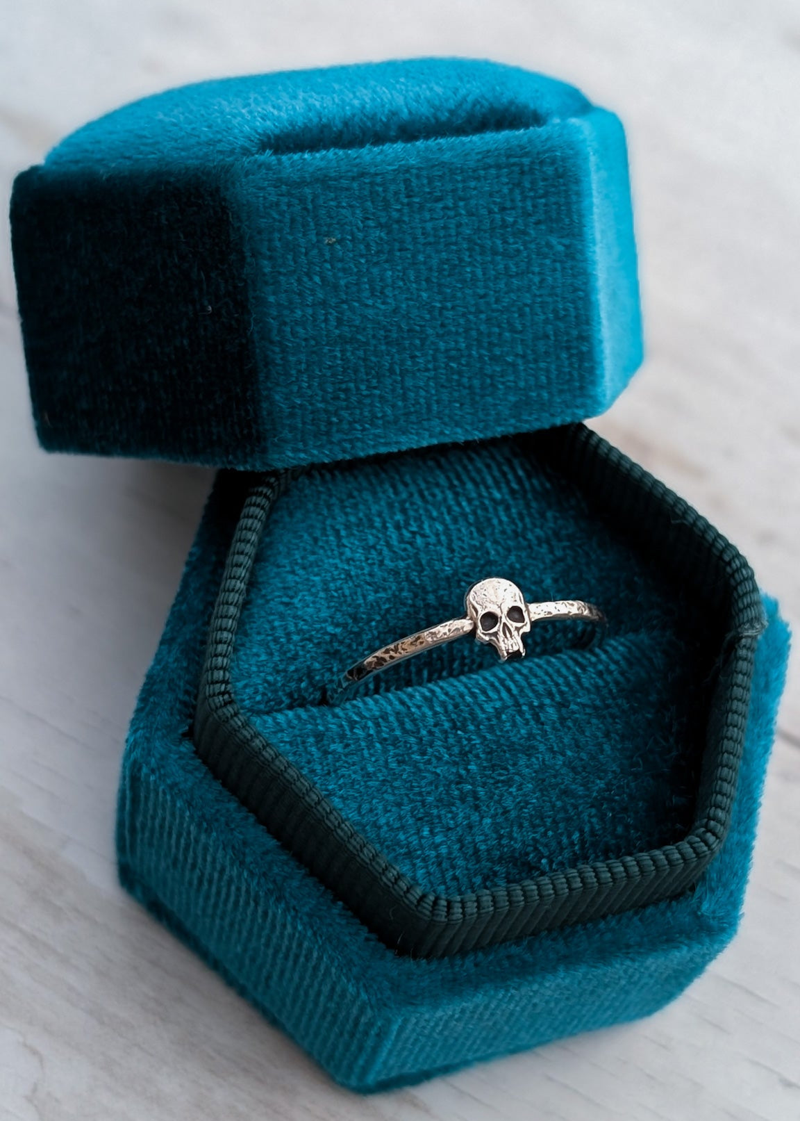 Silver skull ring in a teal blue velvet jewelry box on a light wooden surface