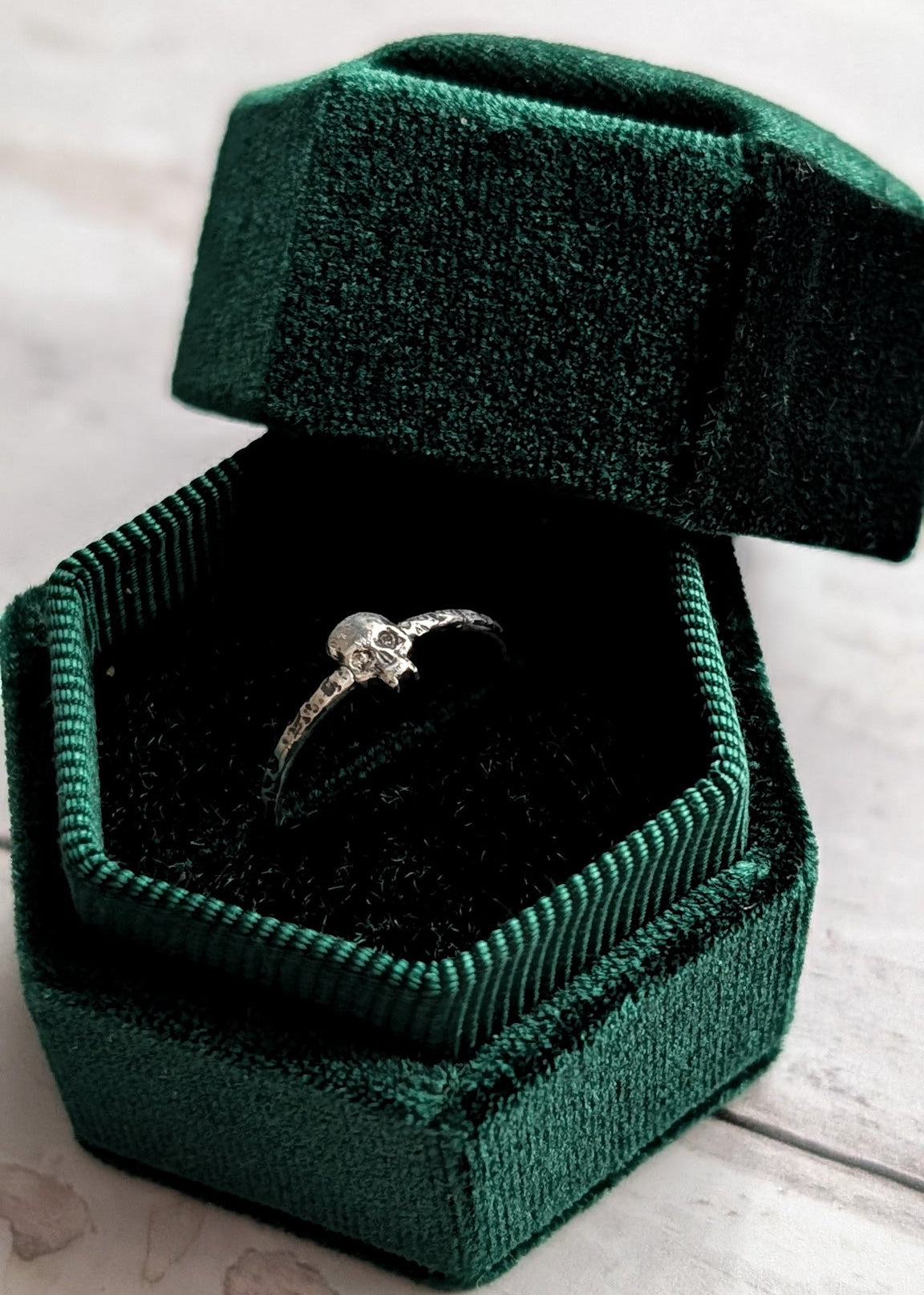 A silver ring with a vampire skull sits inside an open dark green velvet ring box on a light wood surface.