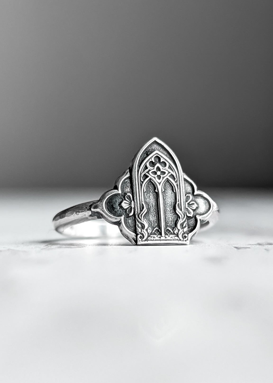 A grey-silver gothic cathedral window ring with floral quatrefoil sides rests on a white surface in front of a warm grey background