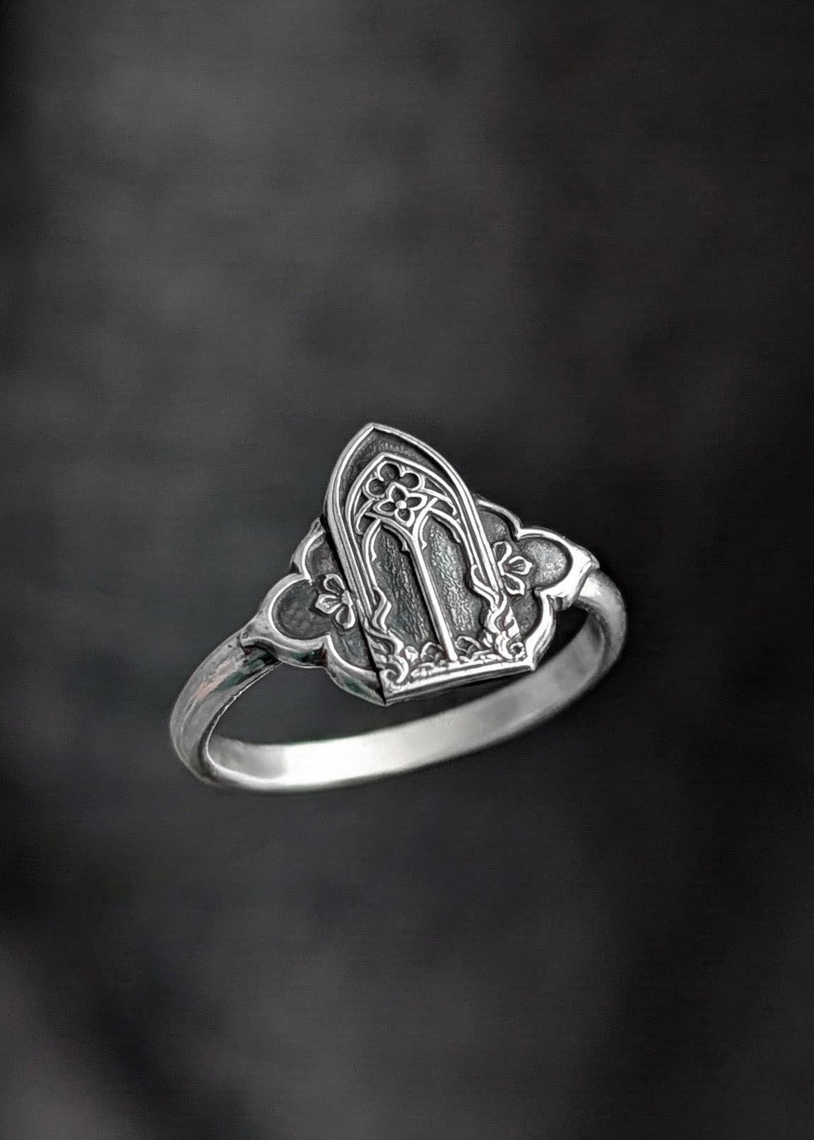 A greyed silver ring with gothic window architecture as the signet emblem floats in front of a soft-focus grey textured background