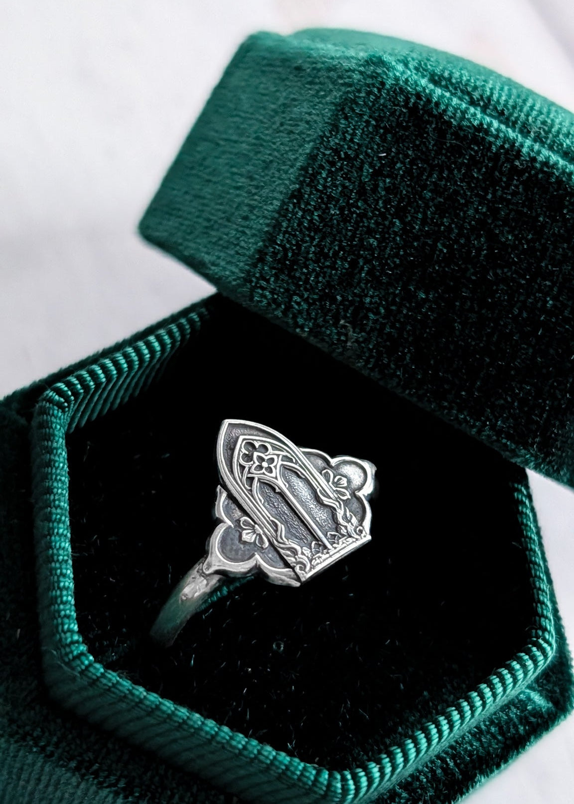 A dark green velvet ring box rests open to present a darkened silver gothic windows saddle ring