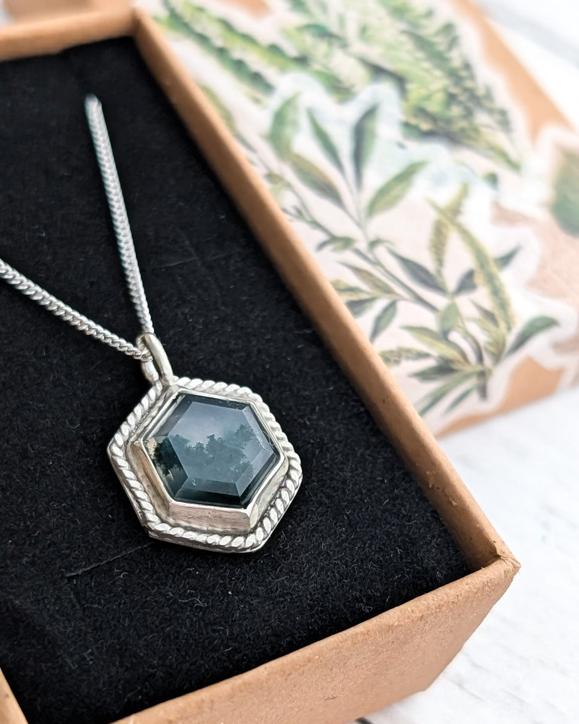 Necklace with a hexagonal mossy gemstone pendant in a gift box with botanical illustration stickers on the lid