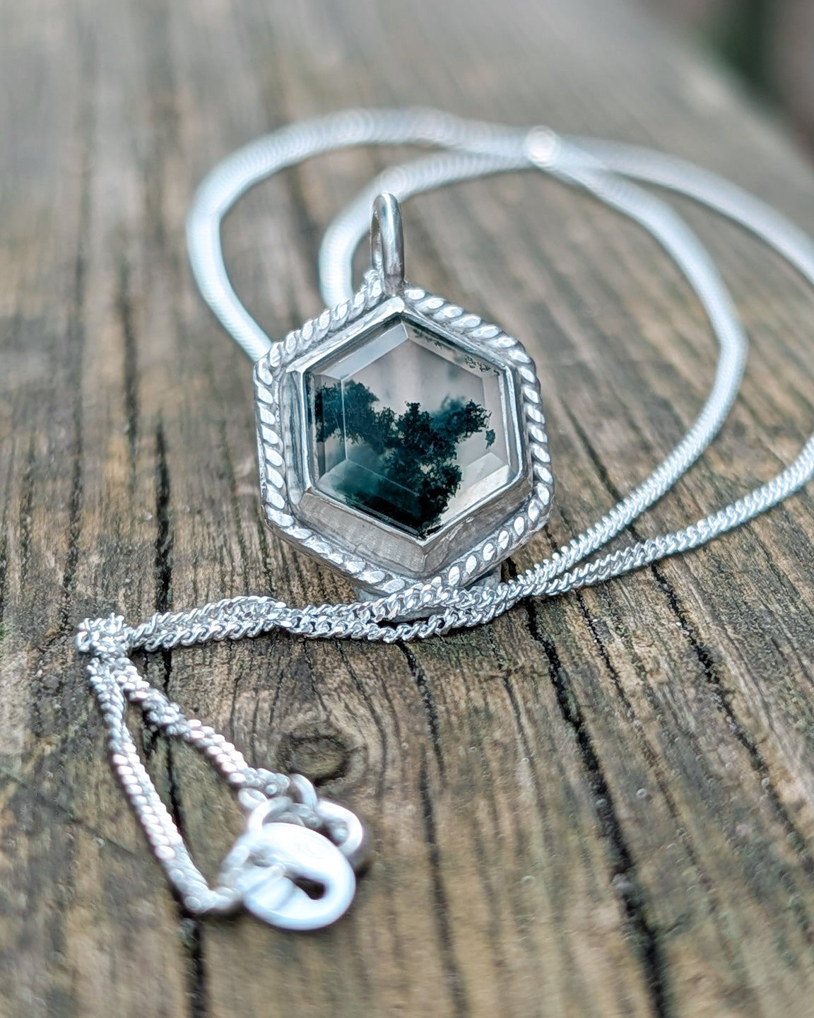 Silver necklace with a hexagonal mossy agate stone pendant resting on a wooden surface