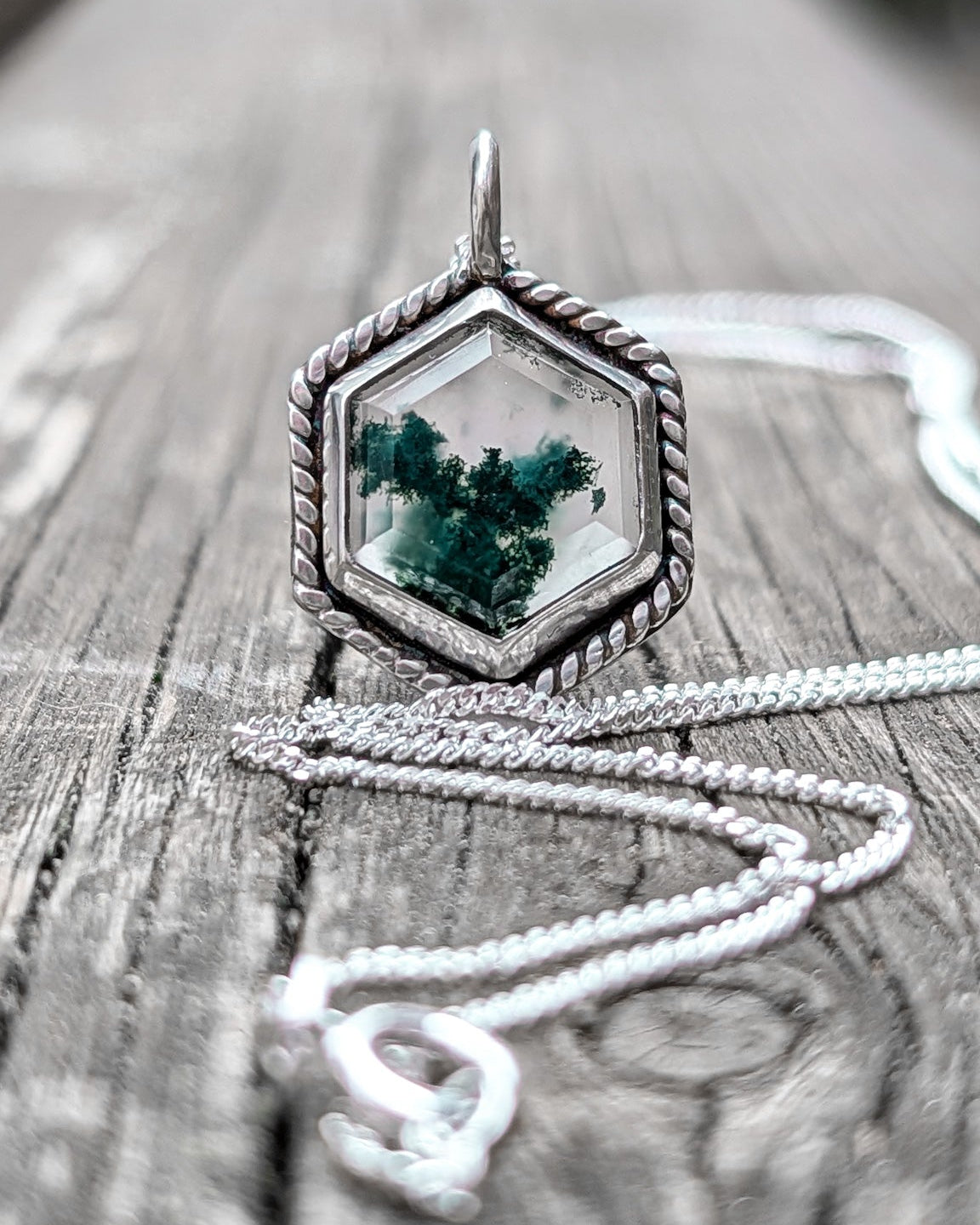 A step cut moss agate hexagon stone is set in a silver pendant with rope wire frame and rests on an old wooden surface.